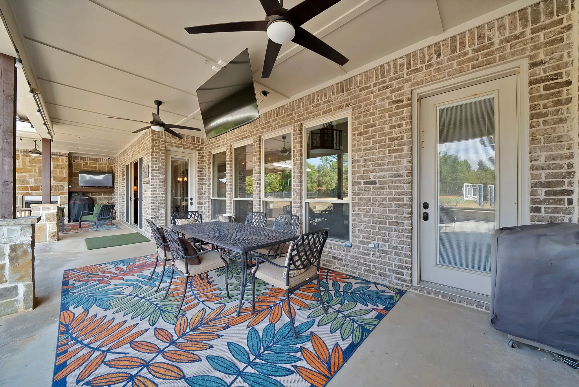 Covered patio with brick and stone exterior, multiple ceiling fans, and an exterior fireplace with a mounted television