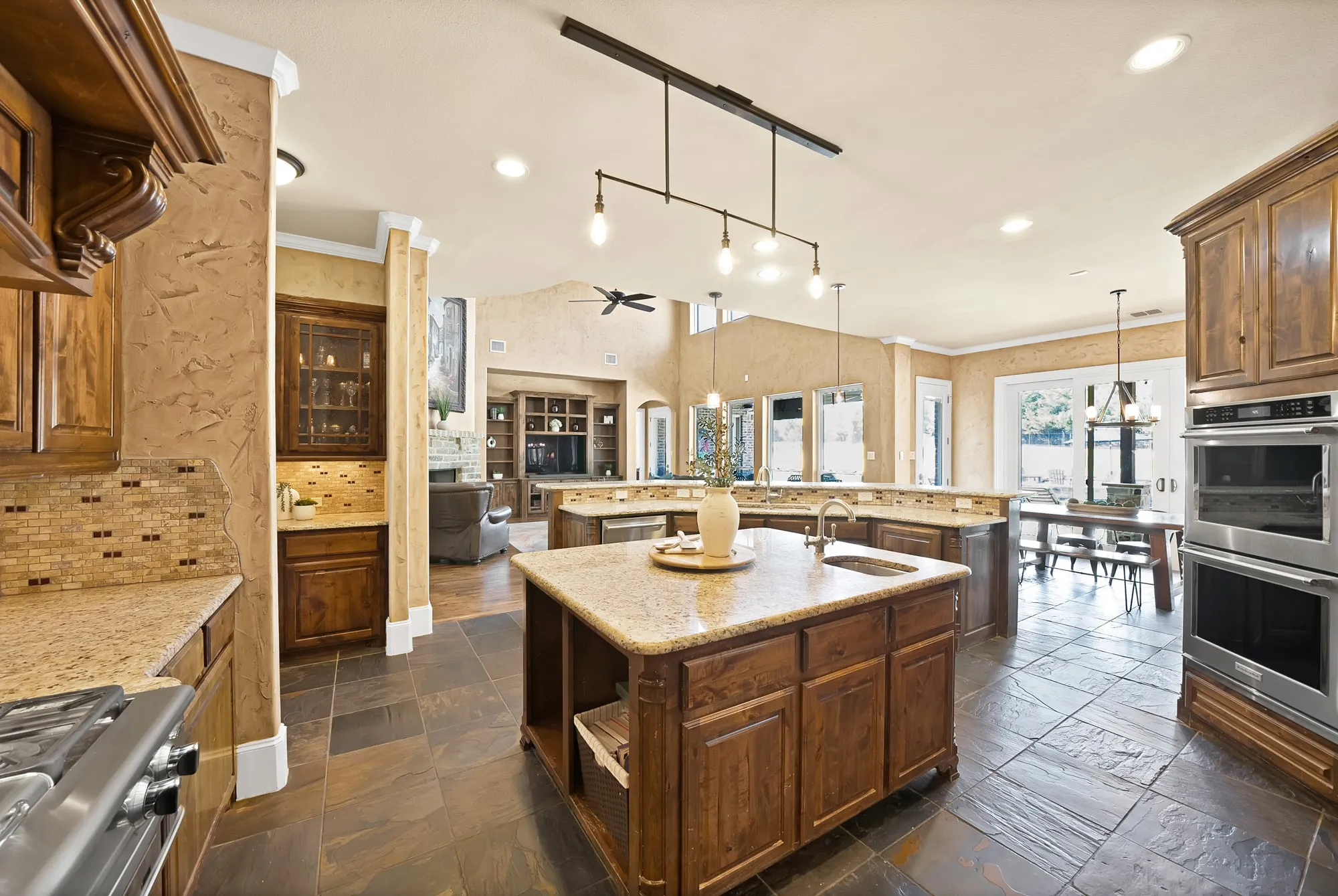 Gourmet kitchen featuring a central island with sink, integrated double ovens, stone tile flooring, and custom wood cabinetry with a mosaic tile backsplash