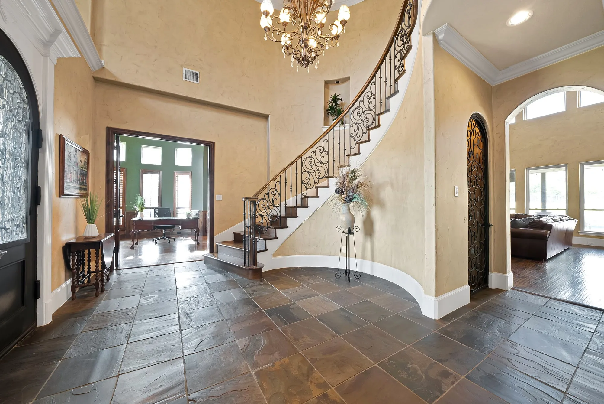 Spacious foyer featuring natural stone flooring, a grand curved staircase with ornate iron balusters, a decorative wrought iron door, and a prominent chandelier