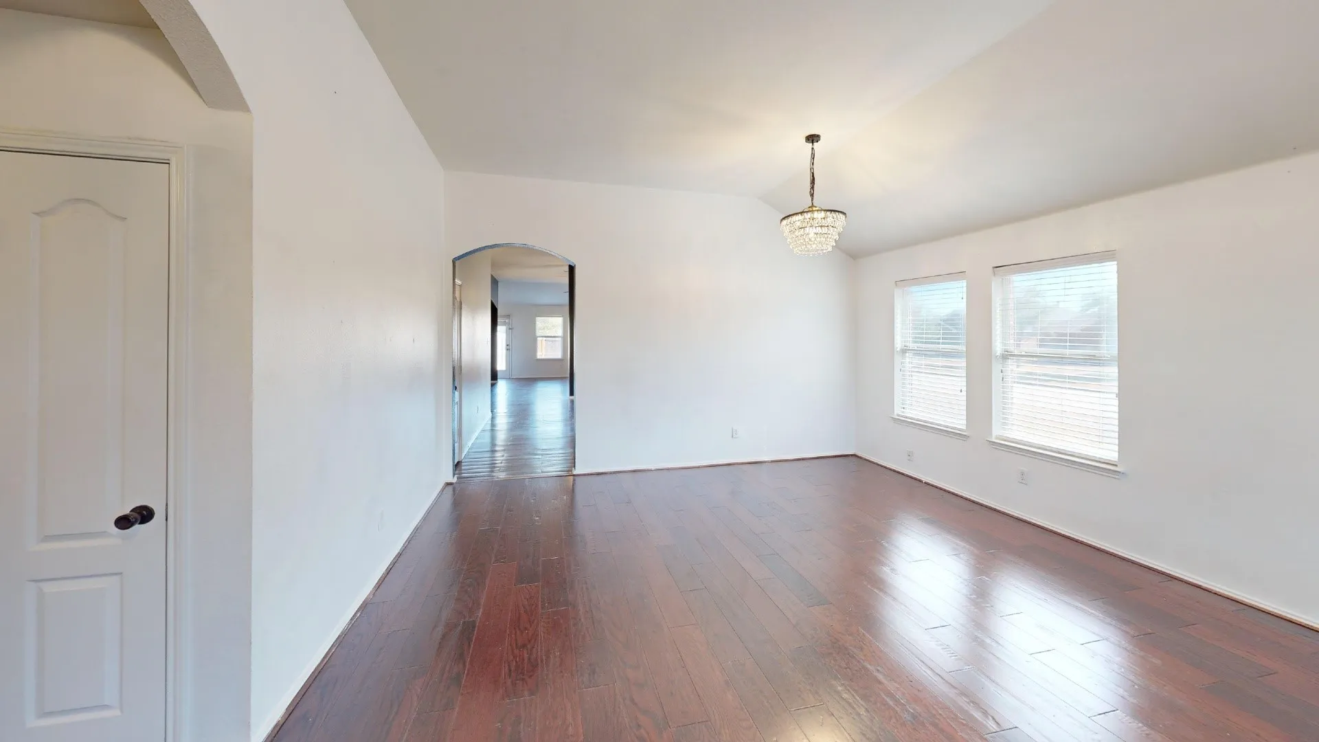 Spacious room featuring wood-finish flooring, vaulted ceiling, dual windows with blinds, and a decorative chandelier