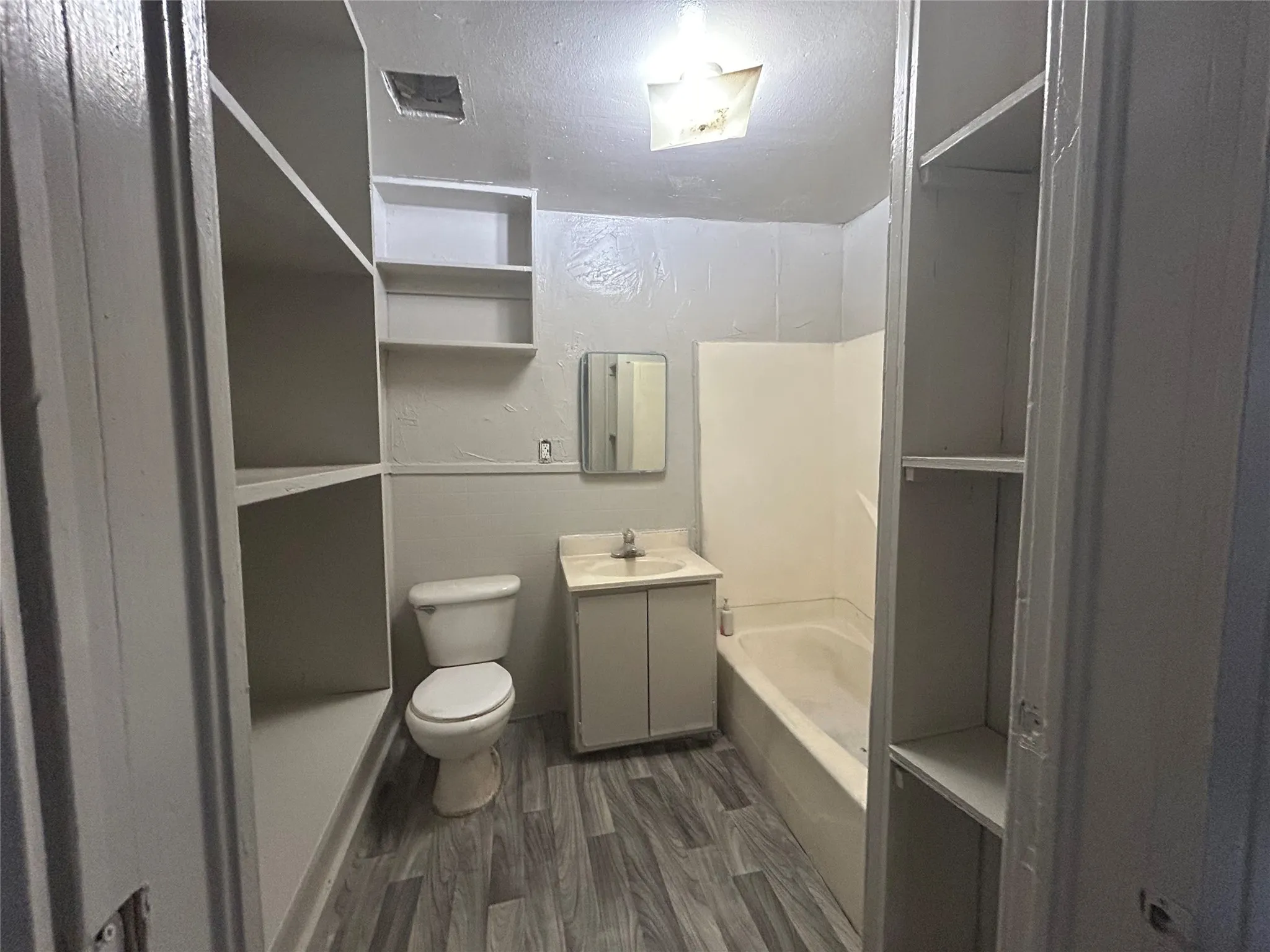 Wood-finish flooring, built-in shelving, white bathtub with a shower surround, vanity with a countertop and sink, and a wall-mounted mirror