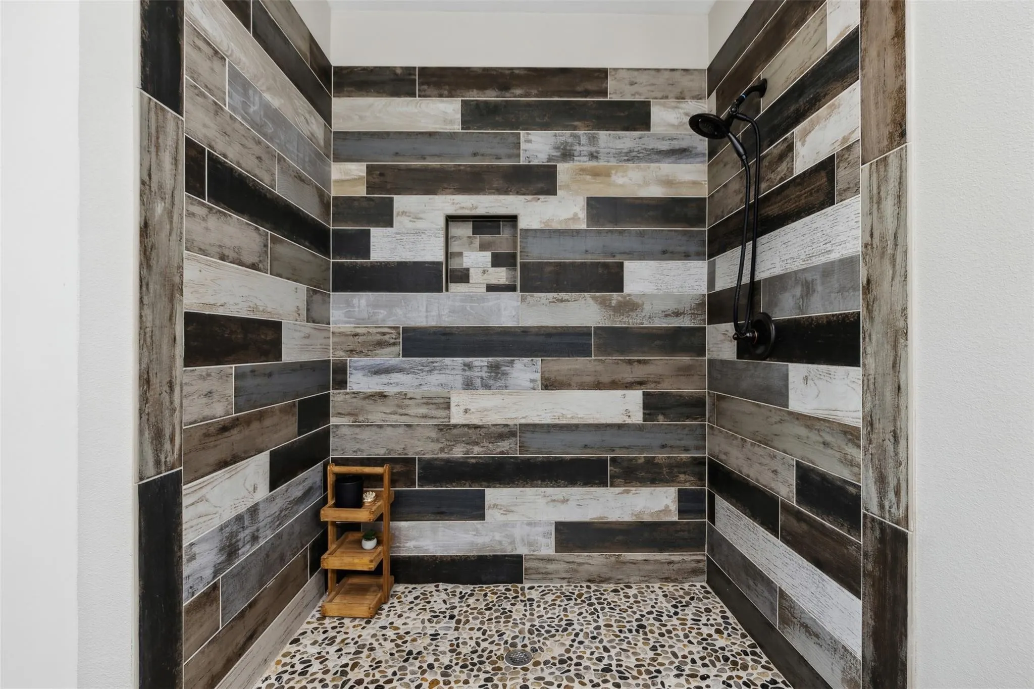 Shower stall featuring wood-finish wall tiling, pebble floor, built-in shower niche, and a matte black shower fixture