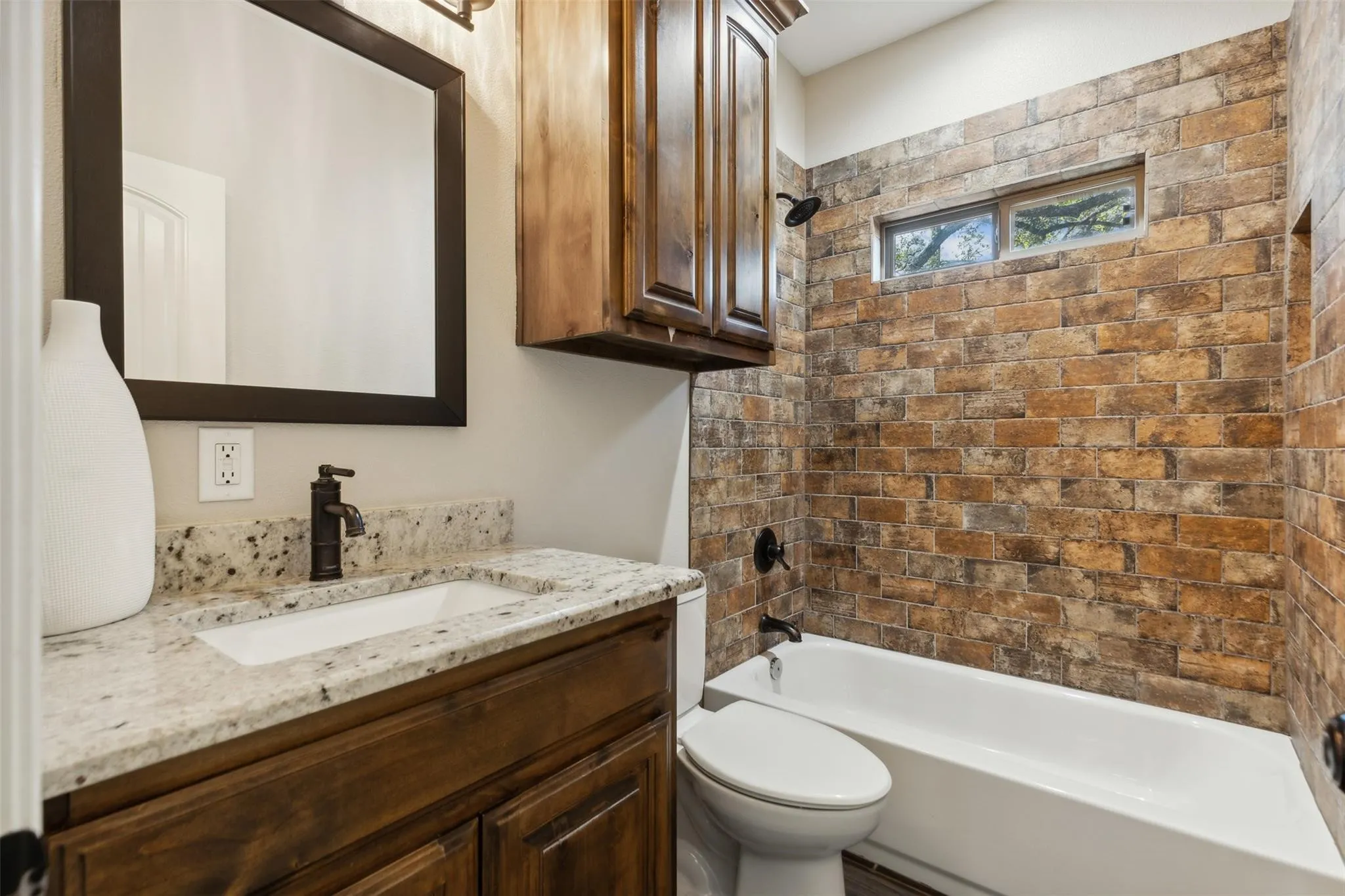 Granite countertop vanity with integrated sink, dark wood-finish cabinetry, framed mirror, and a tiled shower/tub surround