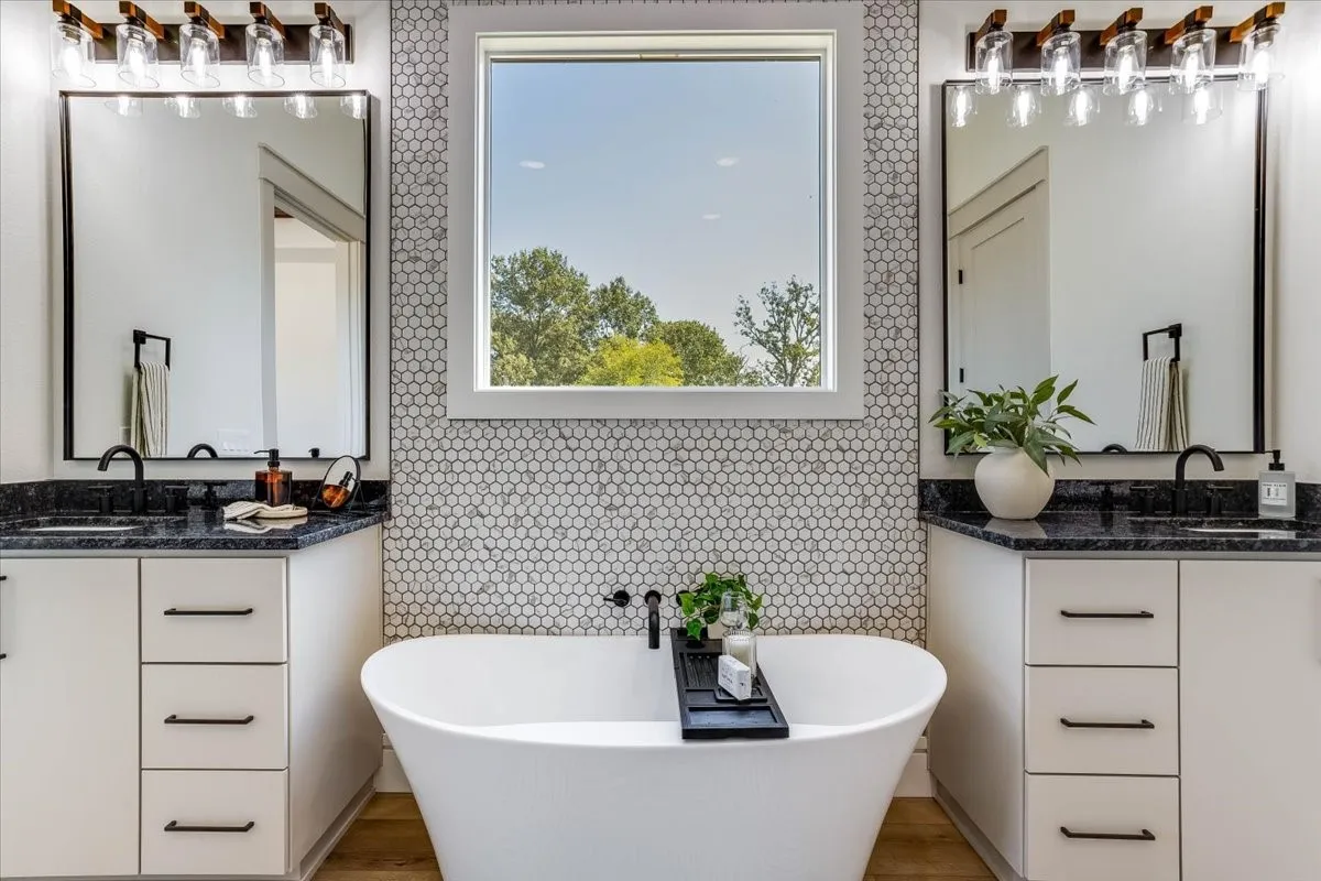 Dual vanities with dark countertops, a large picture window, a freestanding bathtub with a wall-mounted faucet, and honeycomb-patterned wall tile