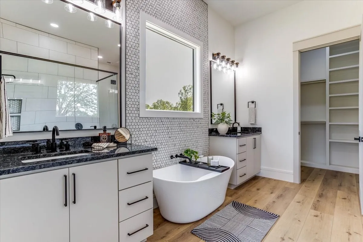 Dual vanities with dark countertops, a freestanding soaking tub, LVP flooring, a mosaic tile accent wall, and built-in closet shelving