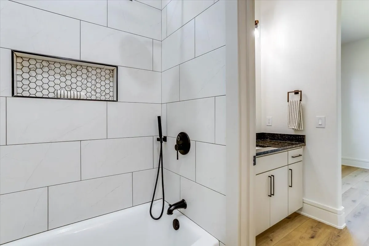 White subway tile surround with integrated hexagonal tile niche, black-finish fixtures, white cabinet vanity with dark countertop, and luxury vinyl plank