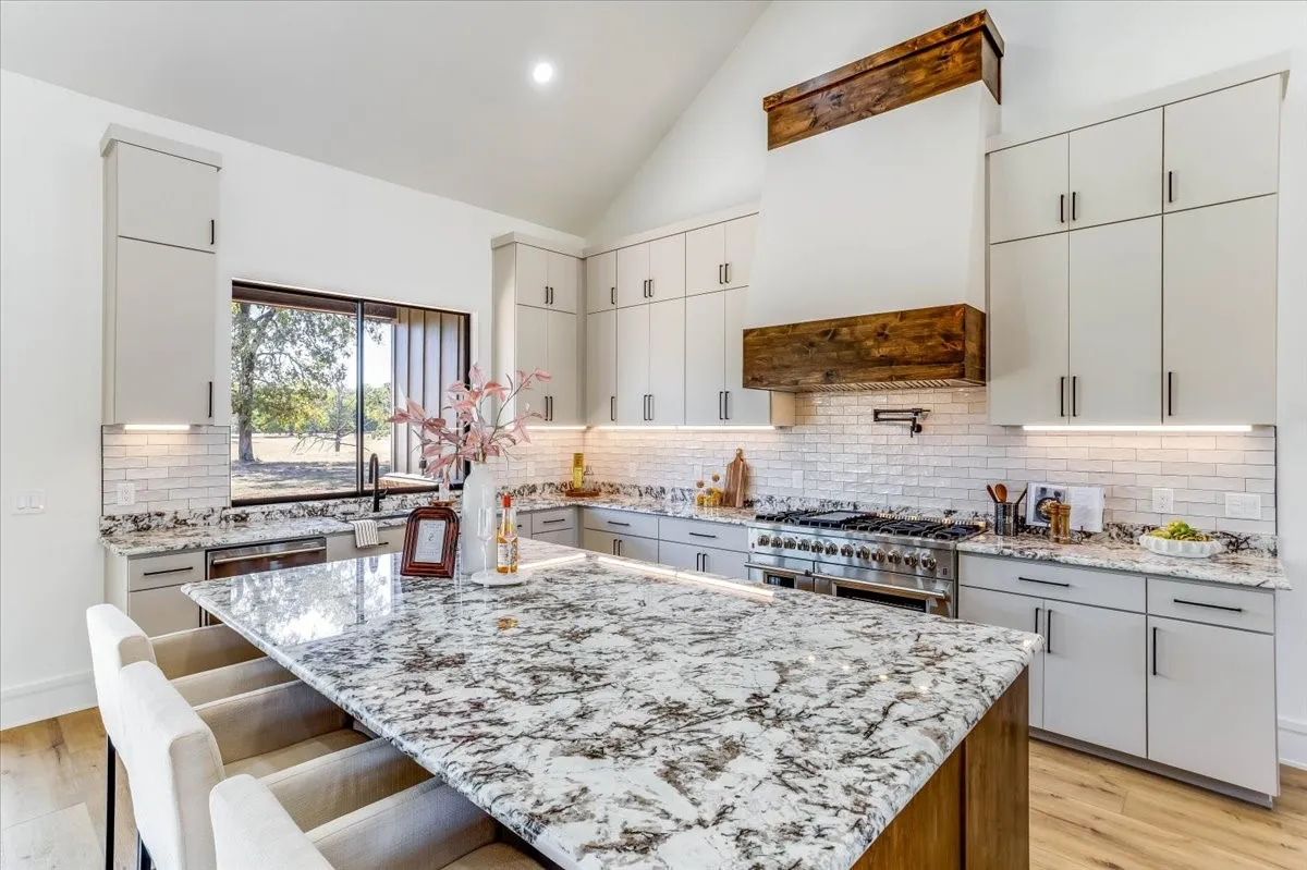 Vaulted ceiling kitchen featuring a large island with a stone countertop, light-toned cabinetry, subway tile backsplash, and stainless steel range