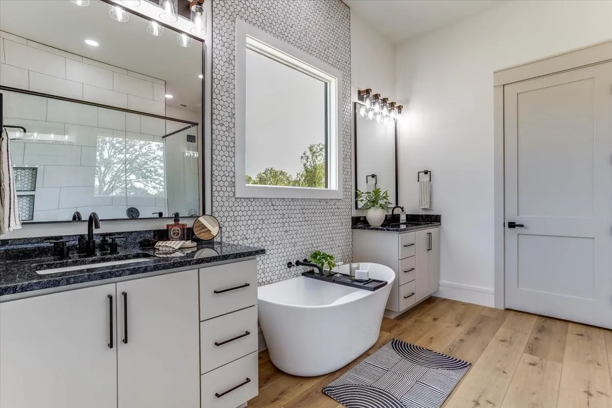Dual vanities with dark countertops, white cabinetry, freestanding soaking tub, LVP flooring, and white penny tile accent wall