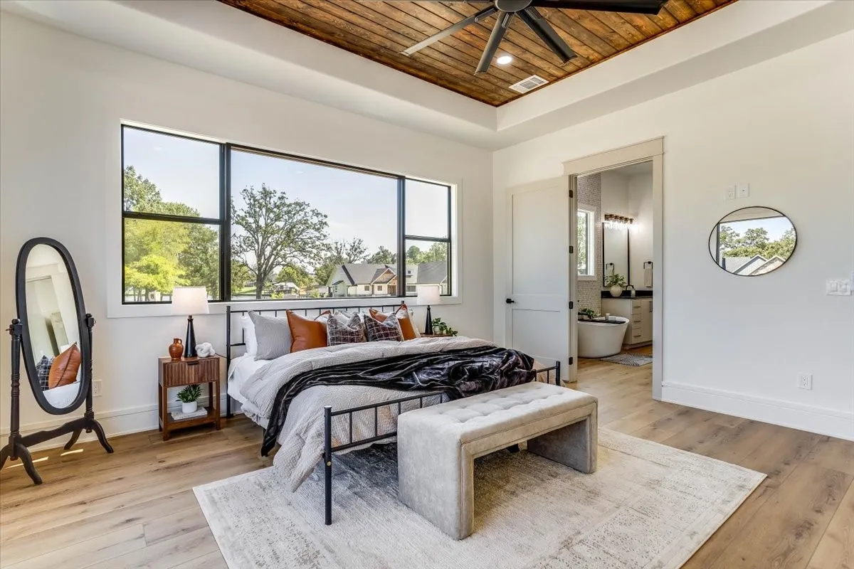 Tray ceiling with wood-finish planks and a ceiling fan, large windows, light wood-finish flooring, and a door leading to a bathroom featuring a freestanding tub and double vanity