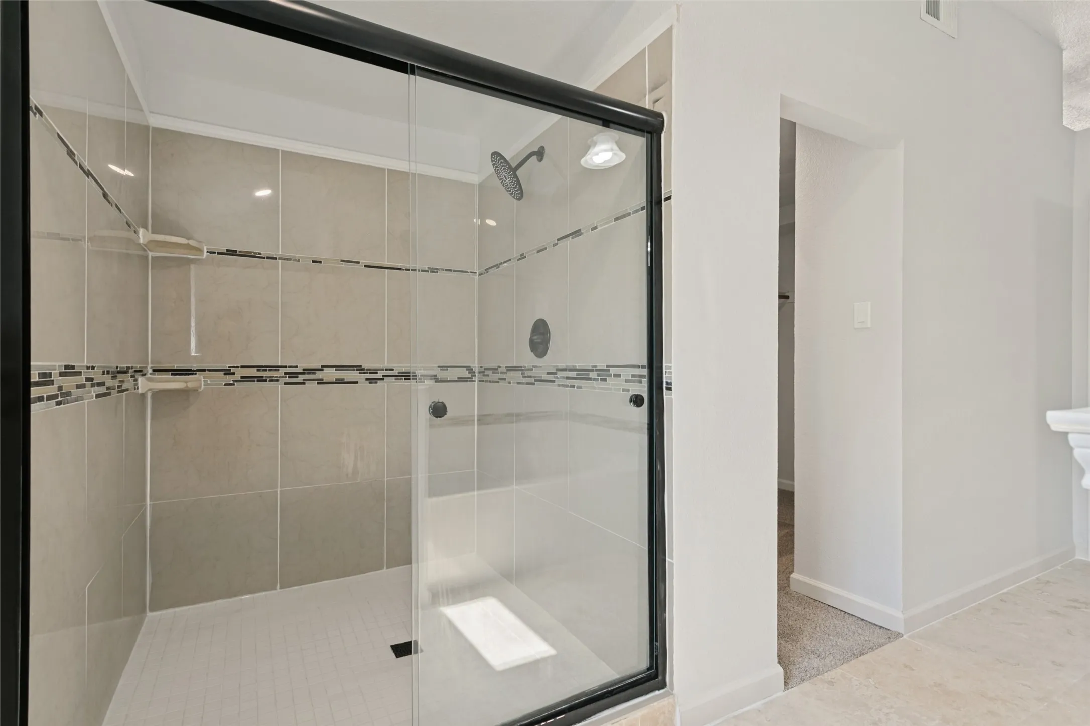 Enclosed shower with clear glass and a black frame, featuring light-toned tile walls, mosaic tile accents, and built-in shower shelves