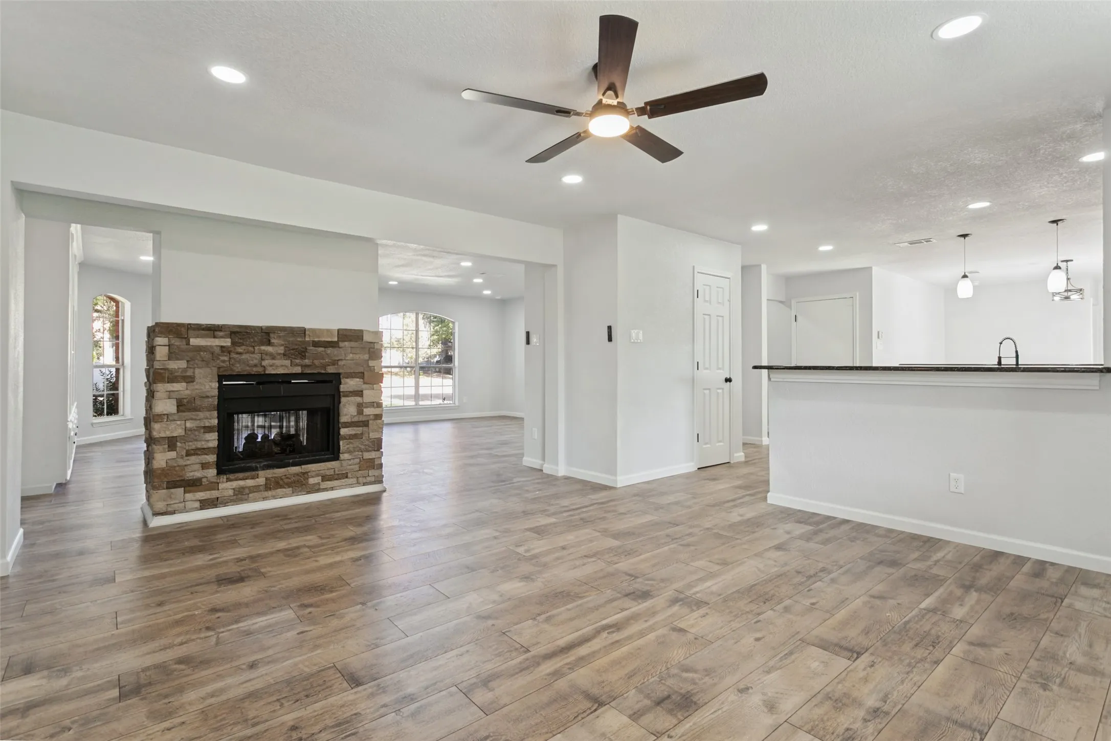 Open-concept living space featuring wood-finish flooring, a stone-clad fireplace, recessed lighting, a ceiling fan, and a breakfast bar