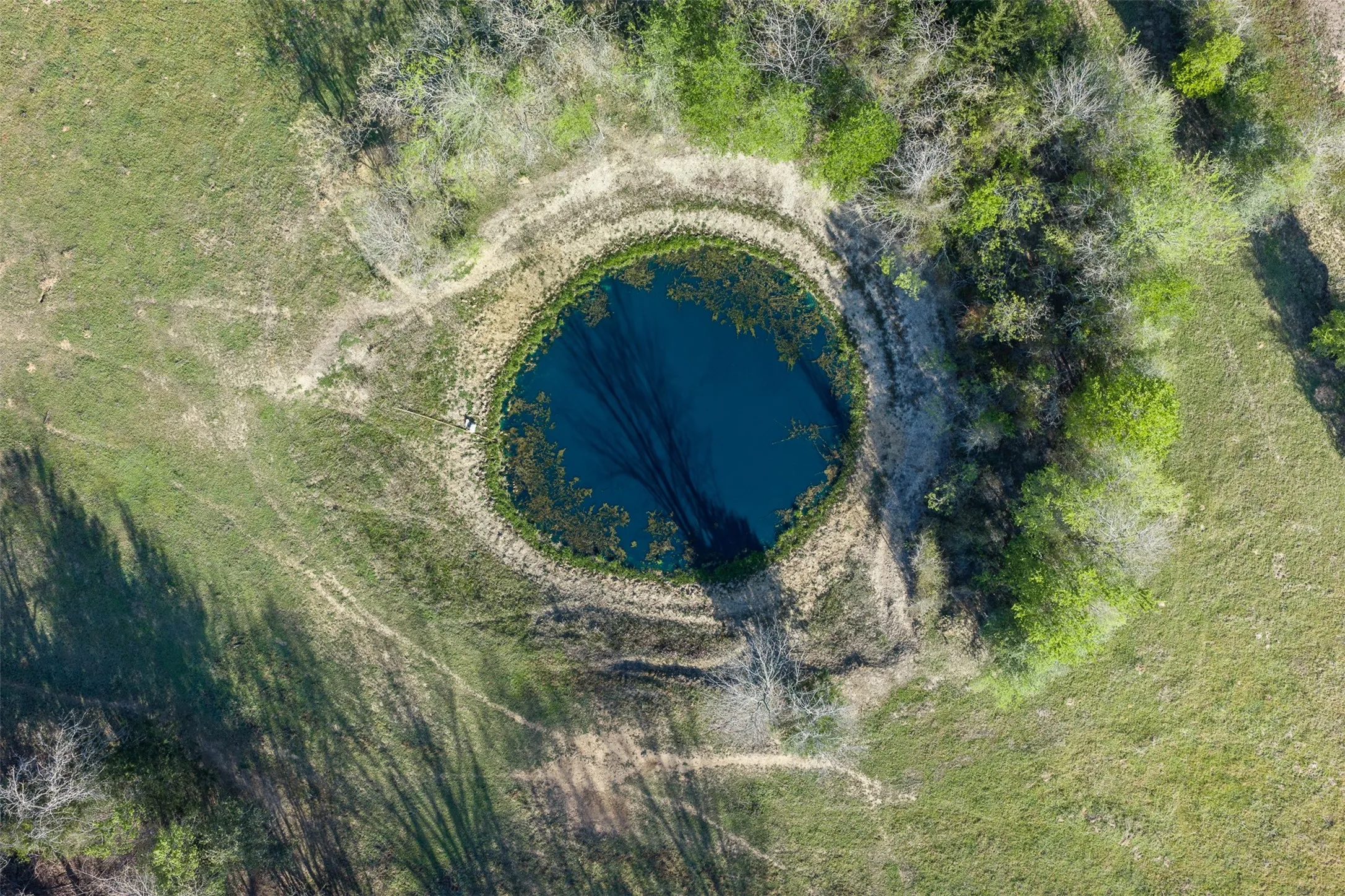 Circular body of water surrounded by vegetation, set within a larger expanse of green and brown terrain
