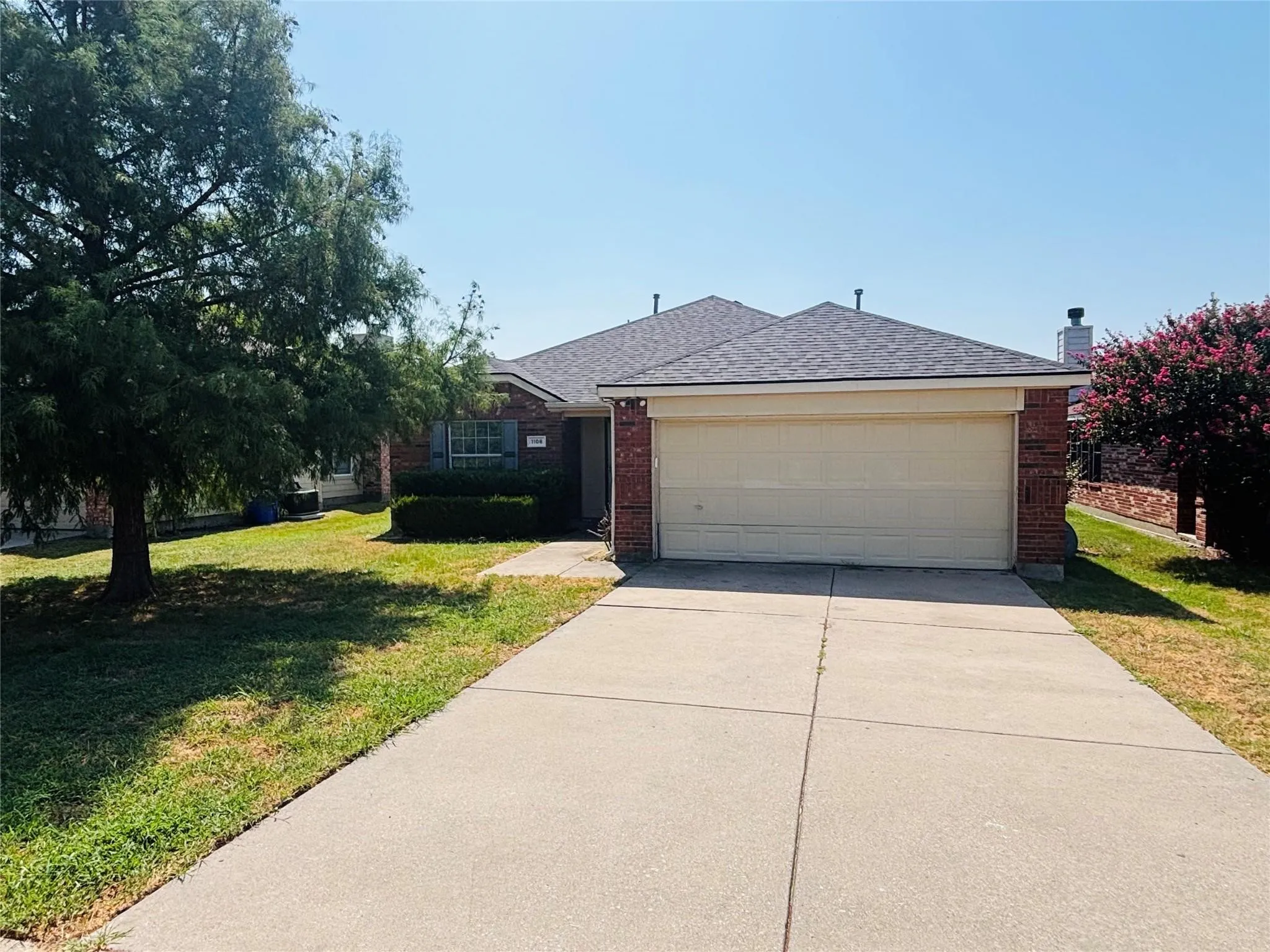 Brick exterior, attached two-car garage, concrete driveway, mature tree, and planted shrubs
