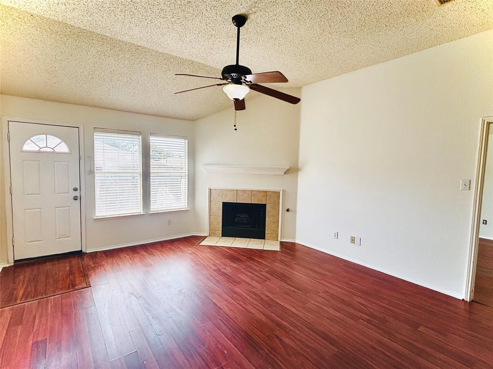 Vaulted ceiling with a ceiling fan, fireplace with a tile surround and mantel, two windows with blinds, front entry door with an arched window, and wood-finish flooring