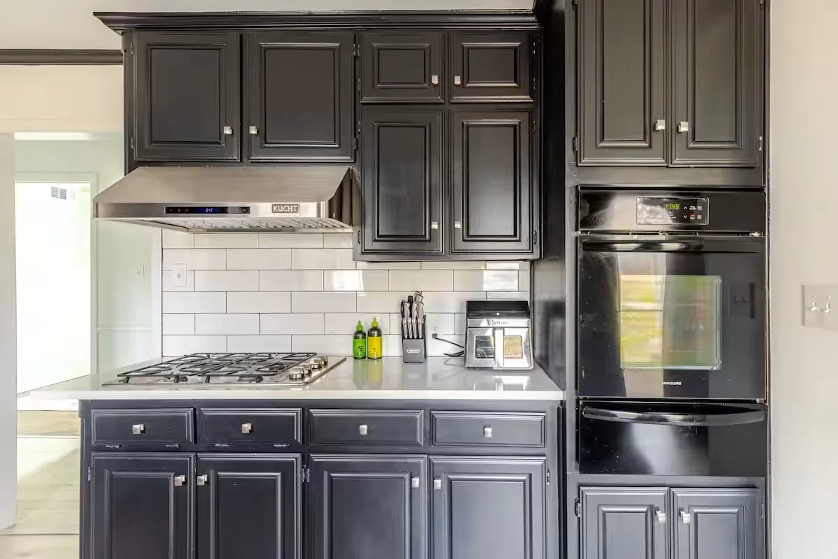 Black cabinetry with panel doors, stainless steel range hood, white subway tile backsplash, white countertop, and built-in wall oven