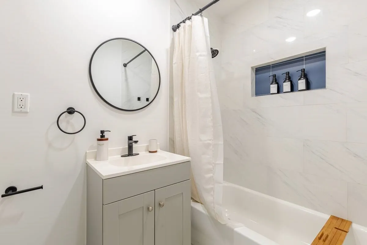 White subway tile shower surround with an integrated niche, matte black fixtures, white bathtub, and a gray vanity with a white countertop