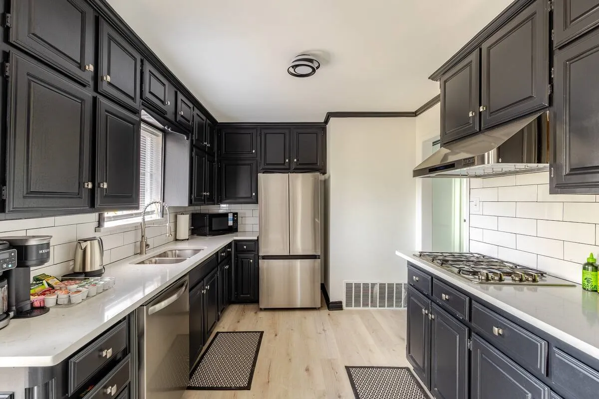 Black shaker-style cabinetry with silver hardware, light wood-finish flooring, stainless steel appliances, white subway tile backsplash, and light-toned countertops