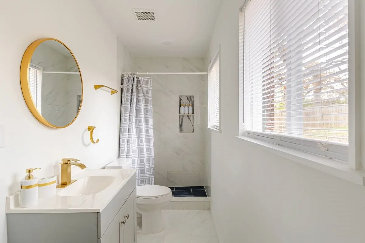 Bathroom featuring a single vanity with gold-finish fixtures, a walk-in shower with tile surround and integrated shelving, white tile flooring, and multiple windows