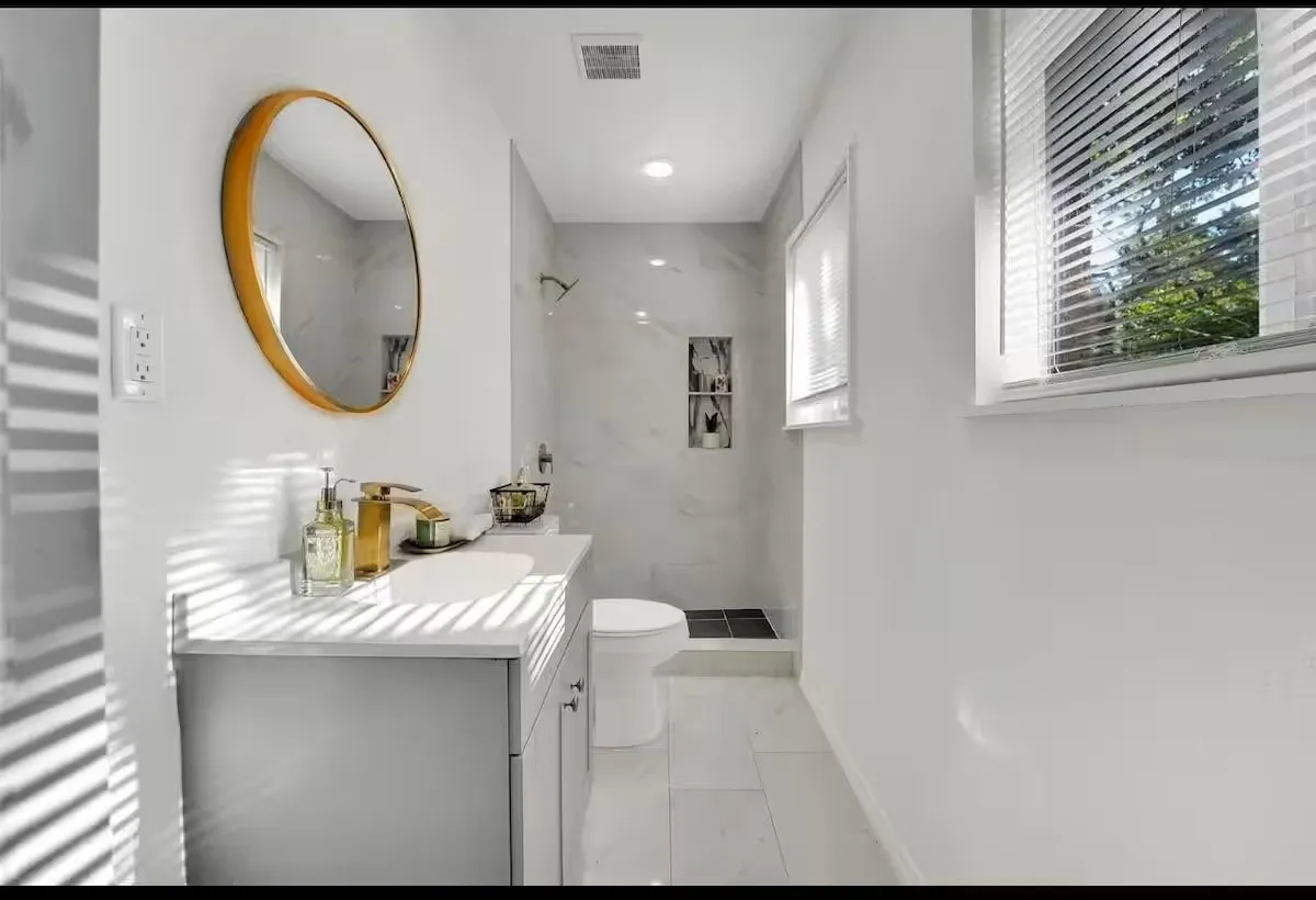 White tiled flooring, vanity with integrated sink, gold-toned faucet, wall-mounted mirror, and tiled shower with built-in niche