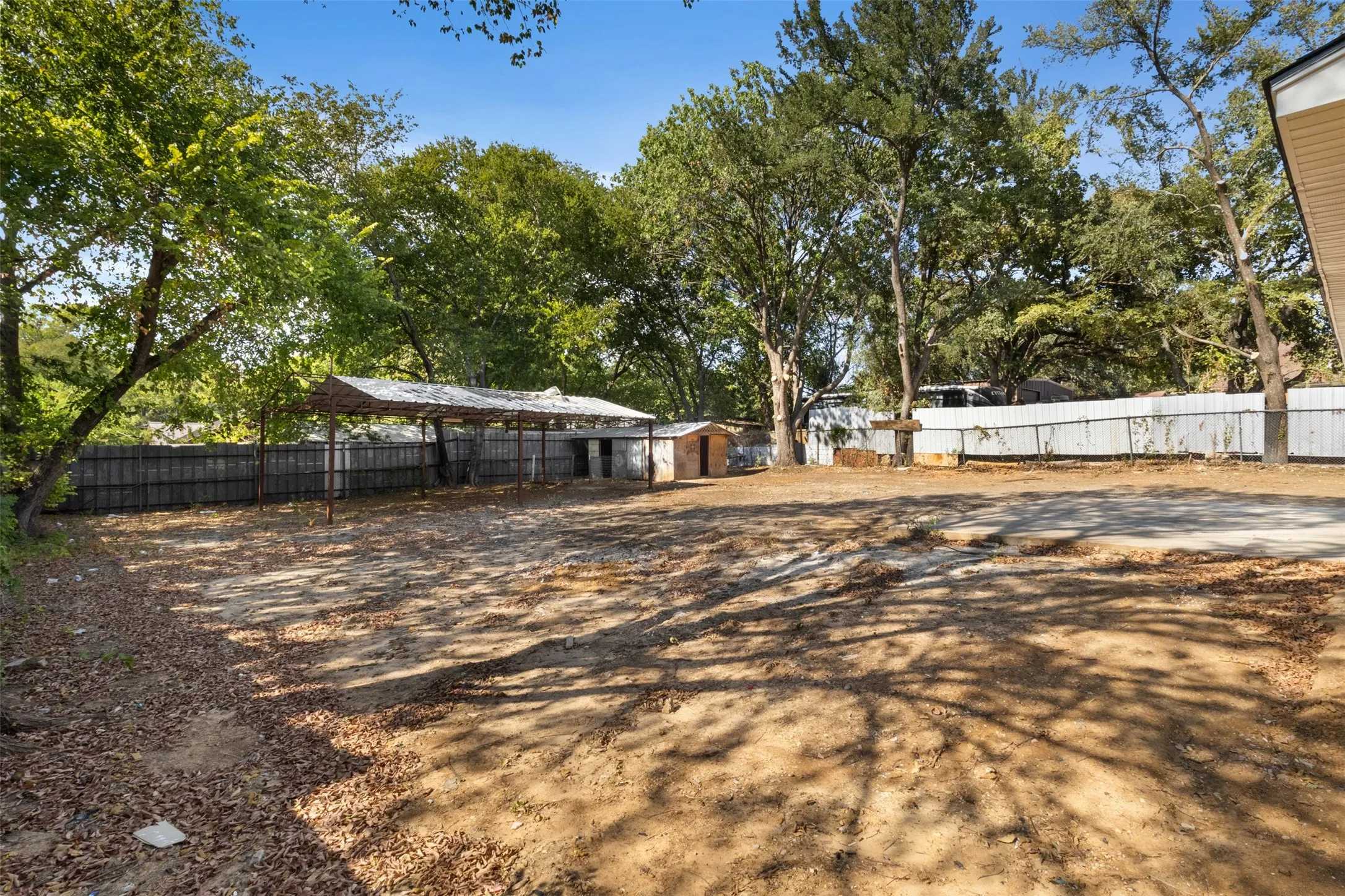 Expansive outdoor area featuring mature trees, a metal-framed covered structure, a utility shed, and perimeter fencing