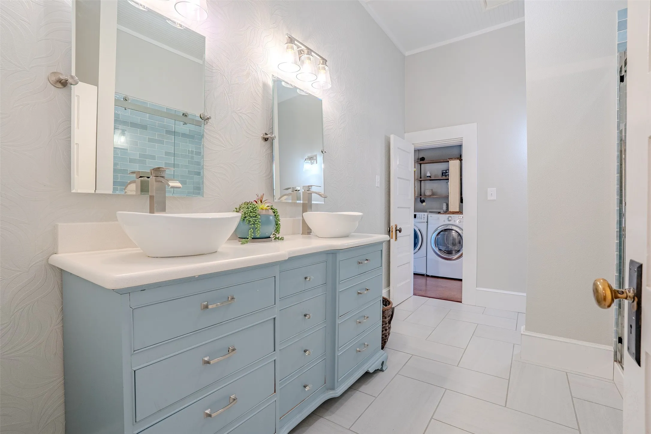 Dual vessel sinks with brushed nickel fixtures, light blue vanity with ample storage, tiled shower with glass door, recessed laundry area with shelving, and light-toned floor tiling