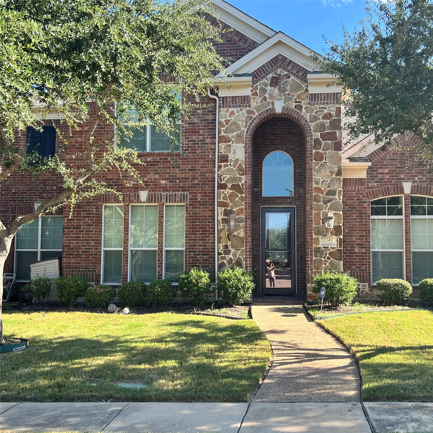 Brick and stone facade, arched entryway, gabled roofline, manicured shrubbery, concrete walkway