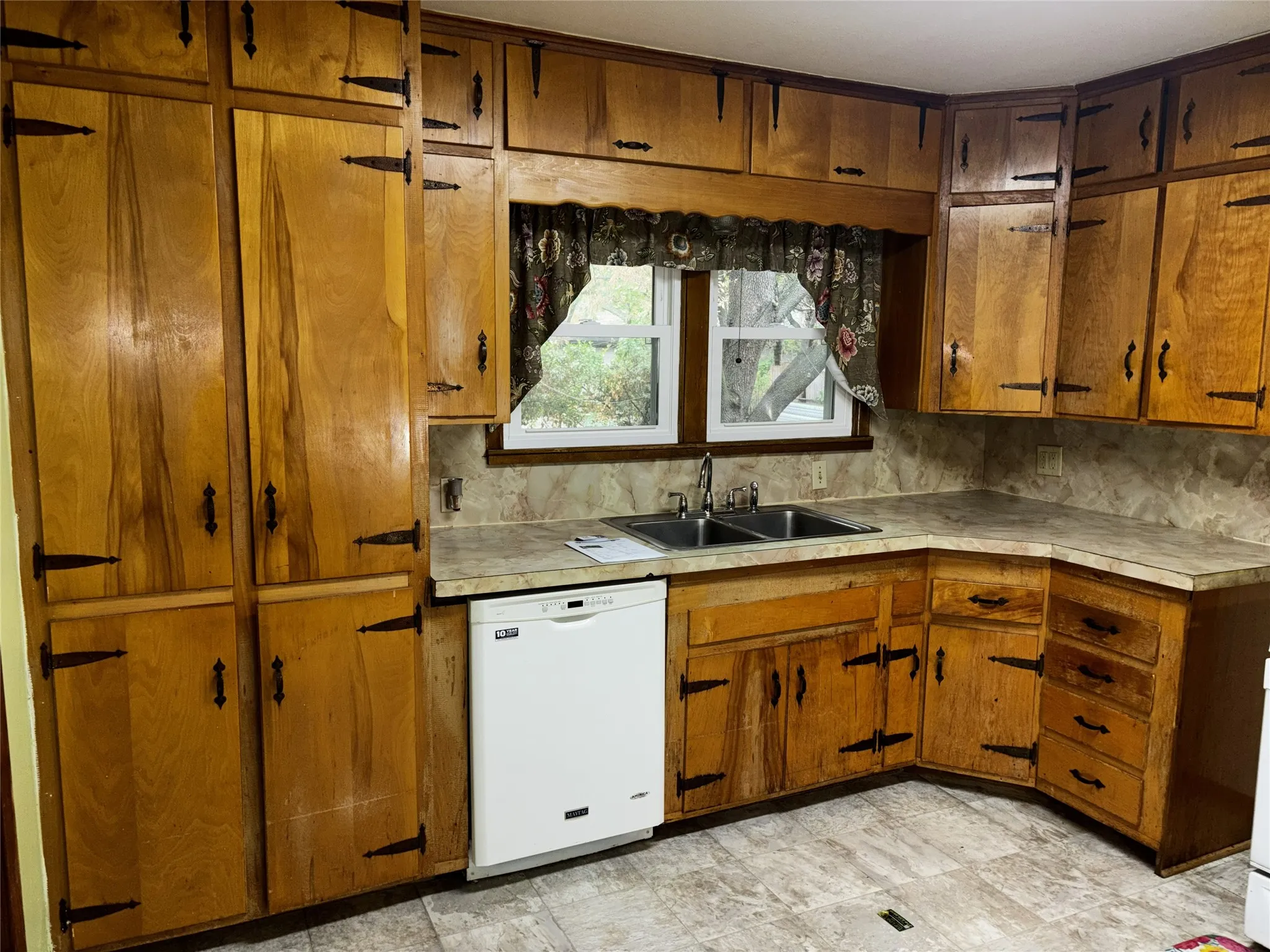 Wood-finish cabinetry with decorative black hardware, L-shaped countertop, double-basin stainless steel sink, and a built-in dishwasher