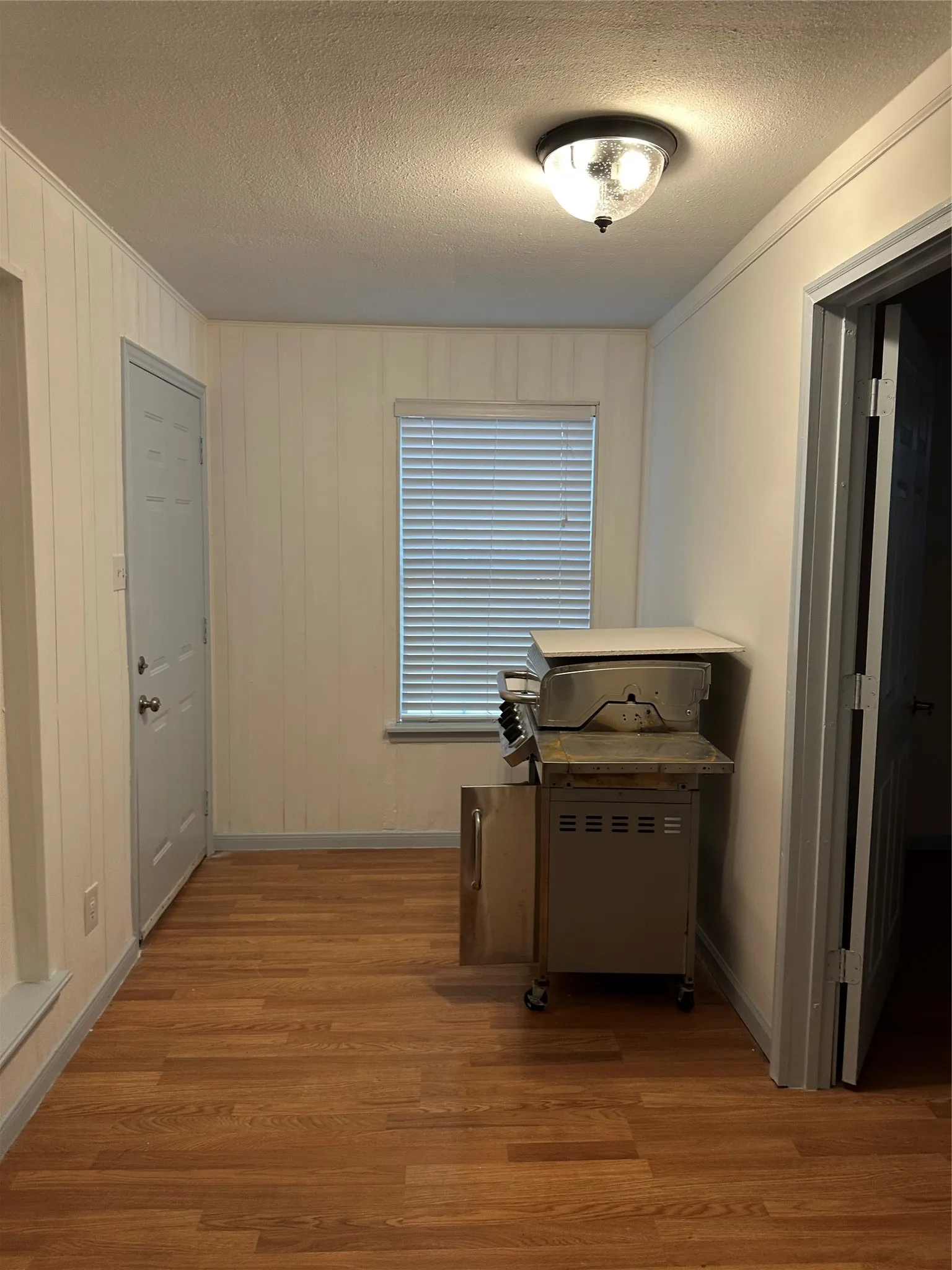 Wood-finish flooring with white paneled walls, a single window with blinds, and a semi-flush mount ceiling light fixture