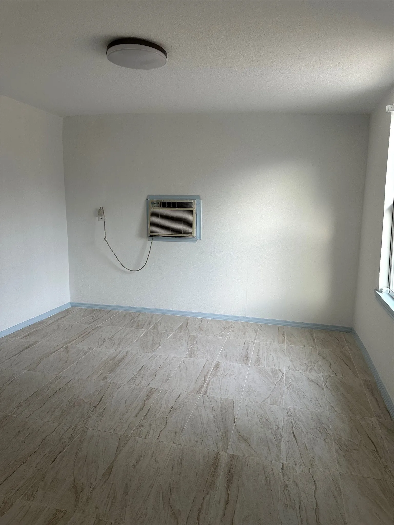 Light-toned tile flooring, white painted walls, window-mounted air conditioning unit, and flush-mount ceiling light