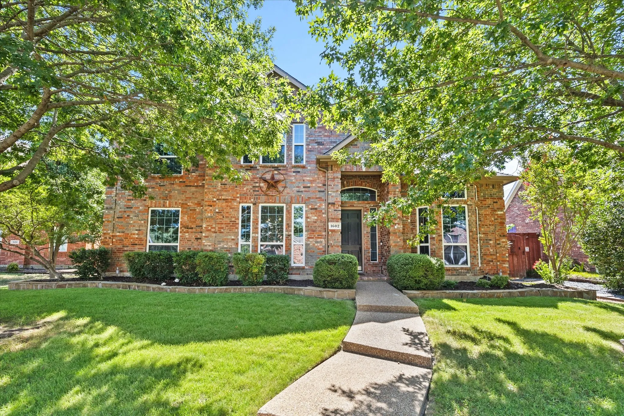 1602 Mapleleaf Drive Wylie, TX 75098- Brick facade with decorative stone accents, mature trees, manicured lawn, established landscaping, and an exposed aggregate walkway