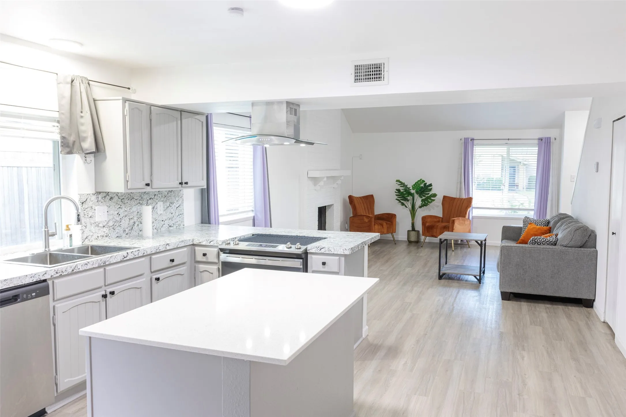 Kitchen with light gray cabinetry, white countertops, and stainless steel appliances, opening to a living area with light wood-finish flooring and a white fireplace surround