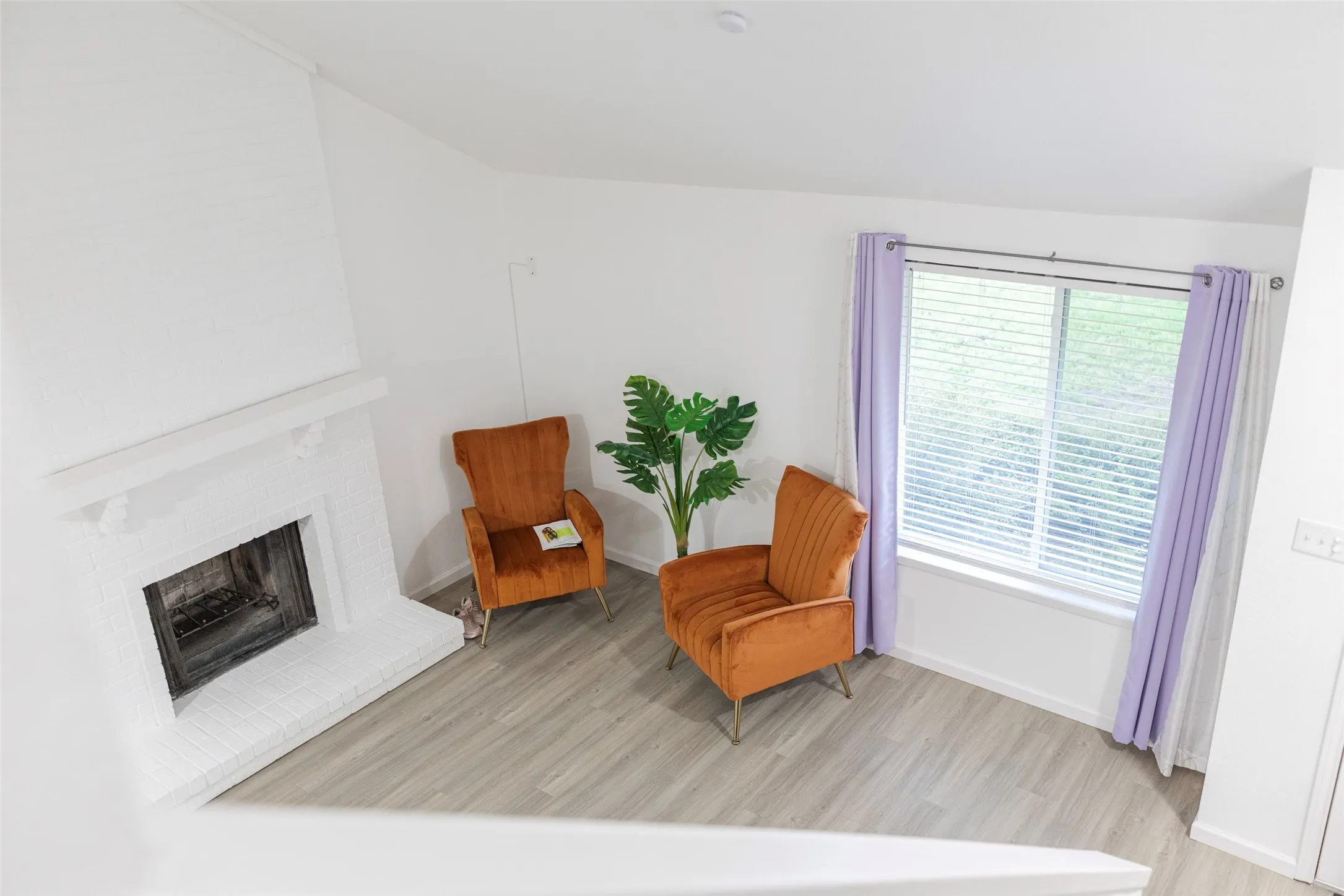 White brick fireplace with mantel, window with blinds, and wood-finish flooring