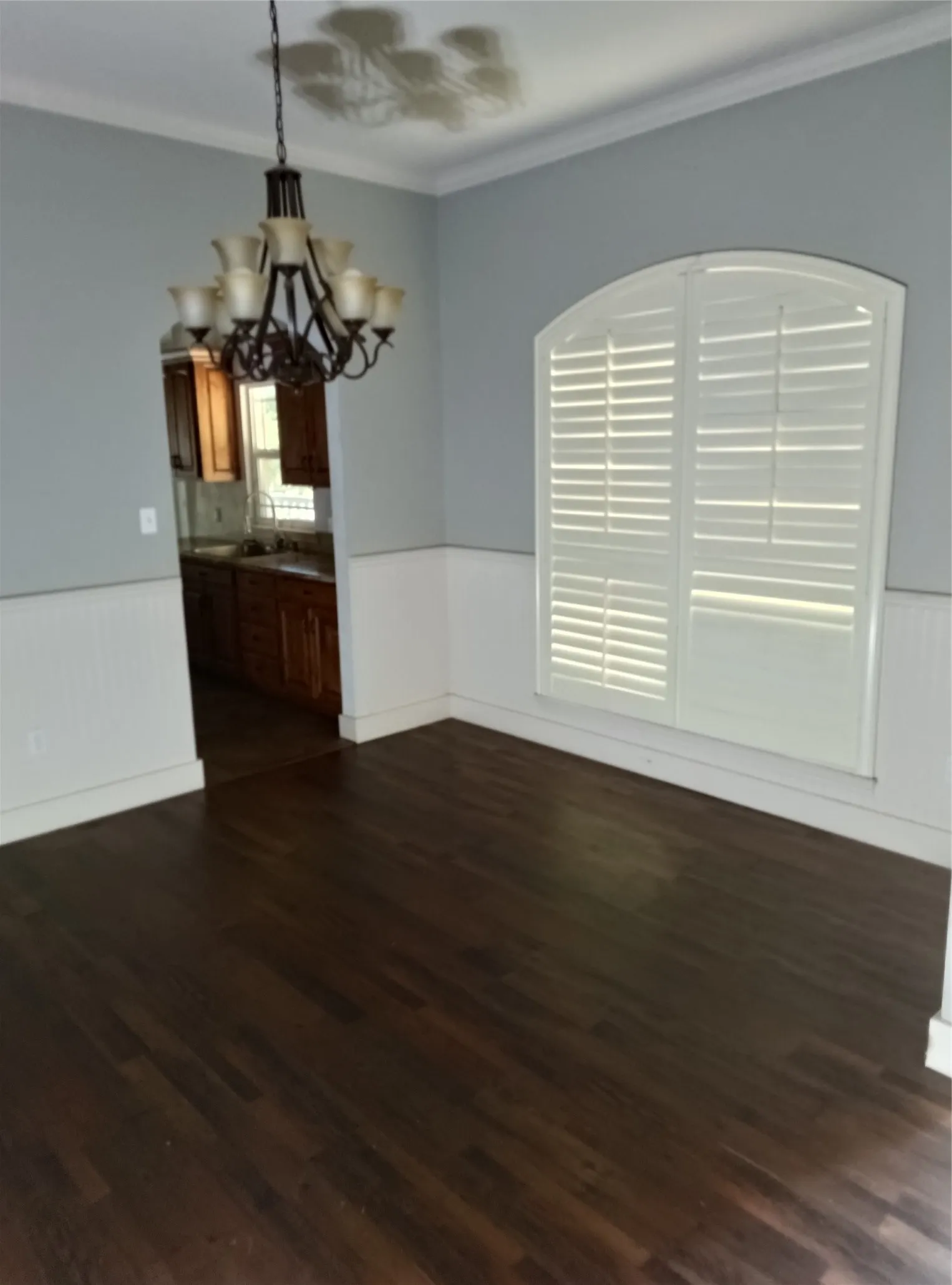 Dark wood-finish flooring, wainscoting, arched window with plantation shutters, crown molding, and a multi-arm chandelier