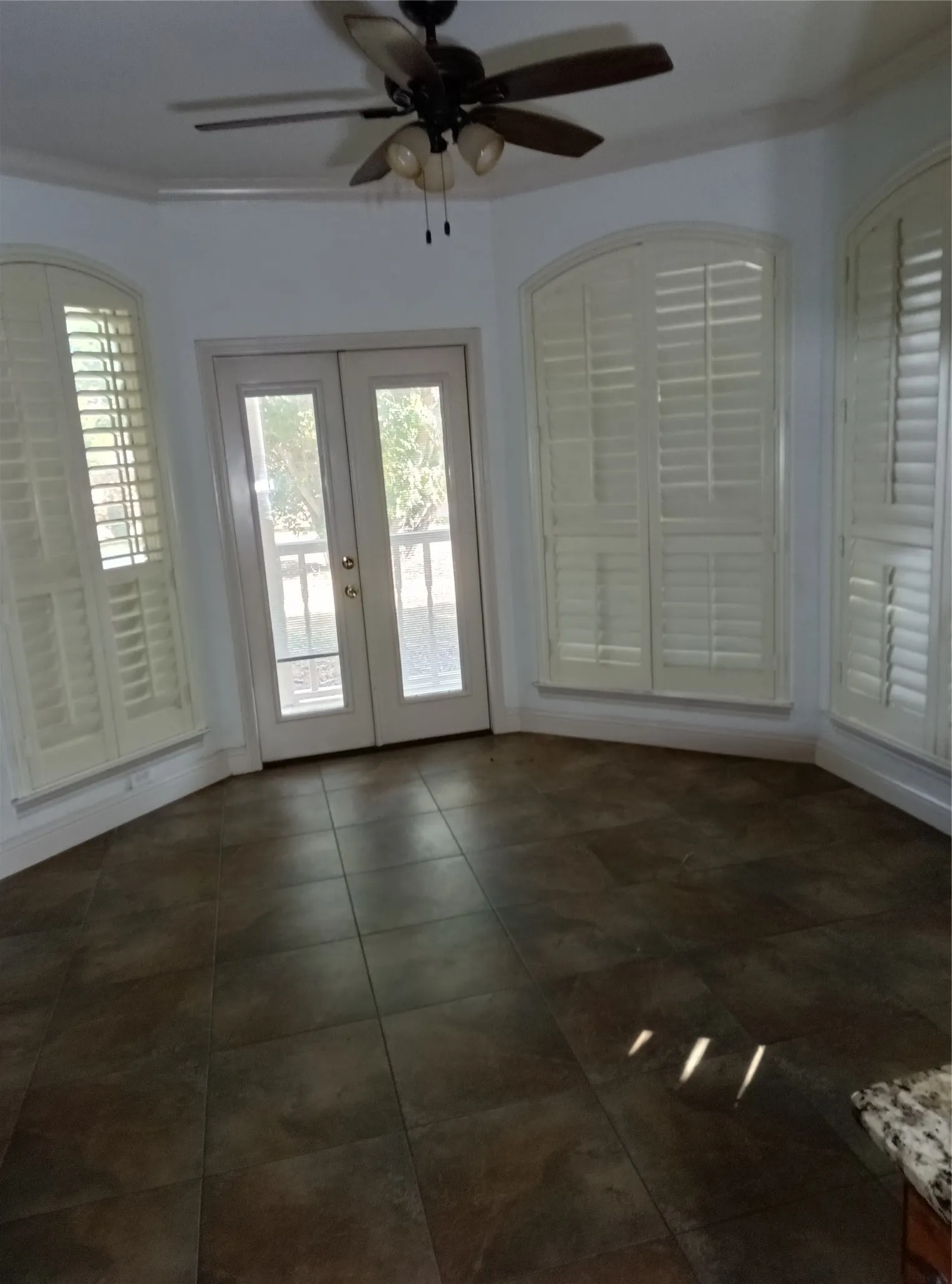 Arched windows with plantation shutters, ceiling fan with integrated lighting, glass-paneled French doors, and square tile flooring