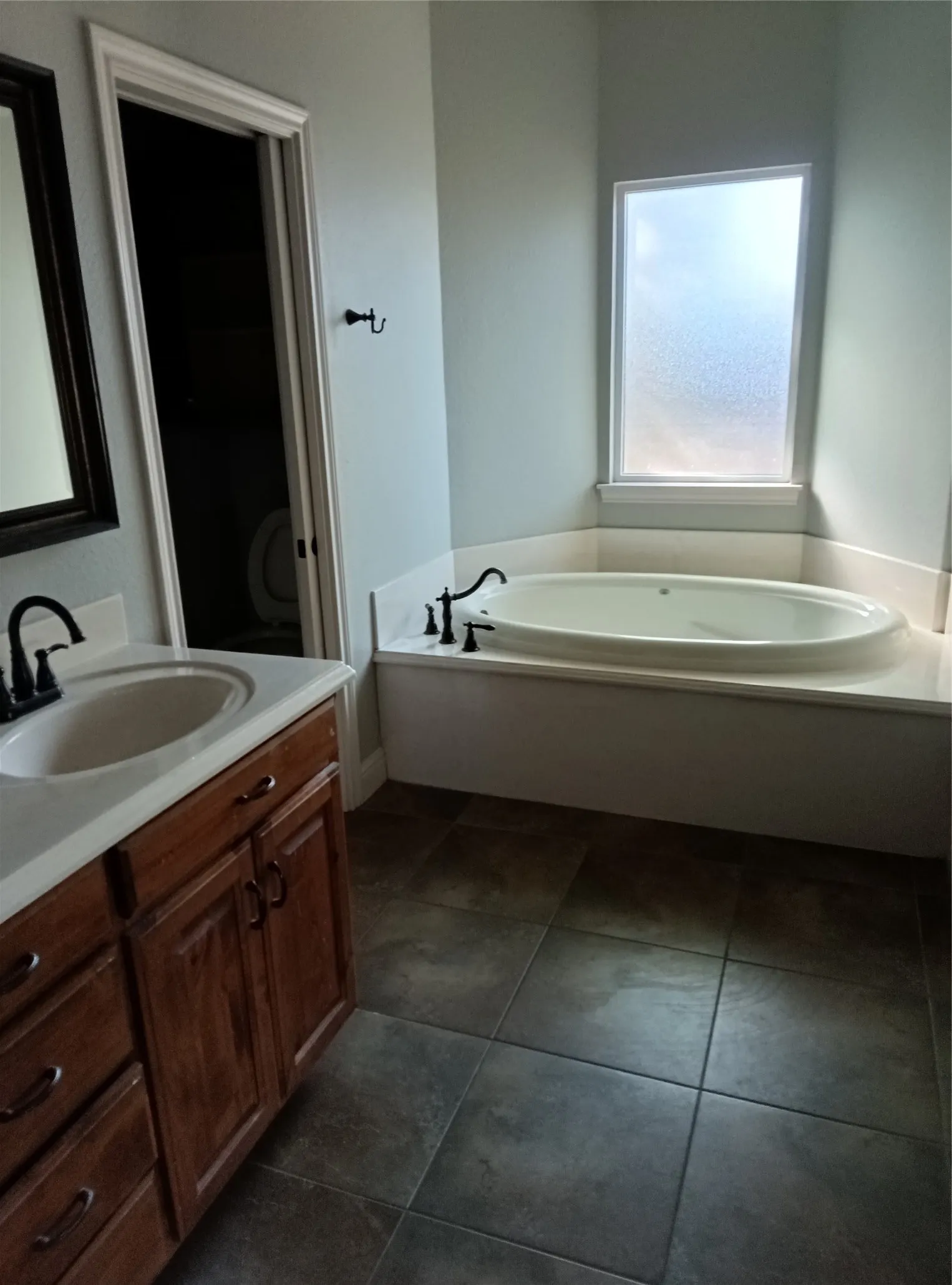 Corner soaking tub with a frosted privacy window, wood-finish vanity with an integrated sink, and dark tiled flooring