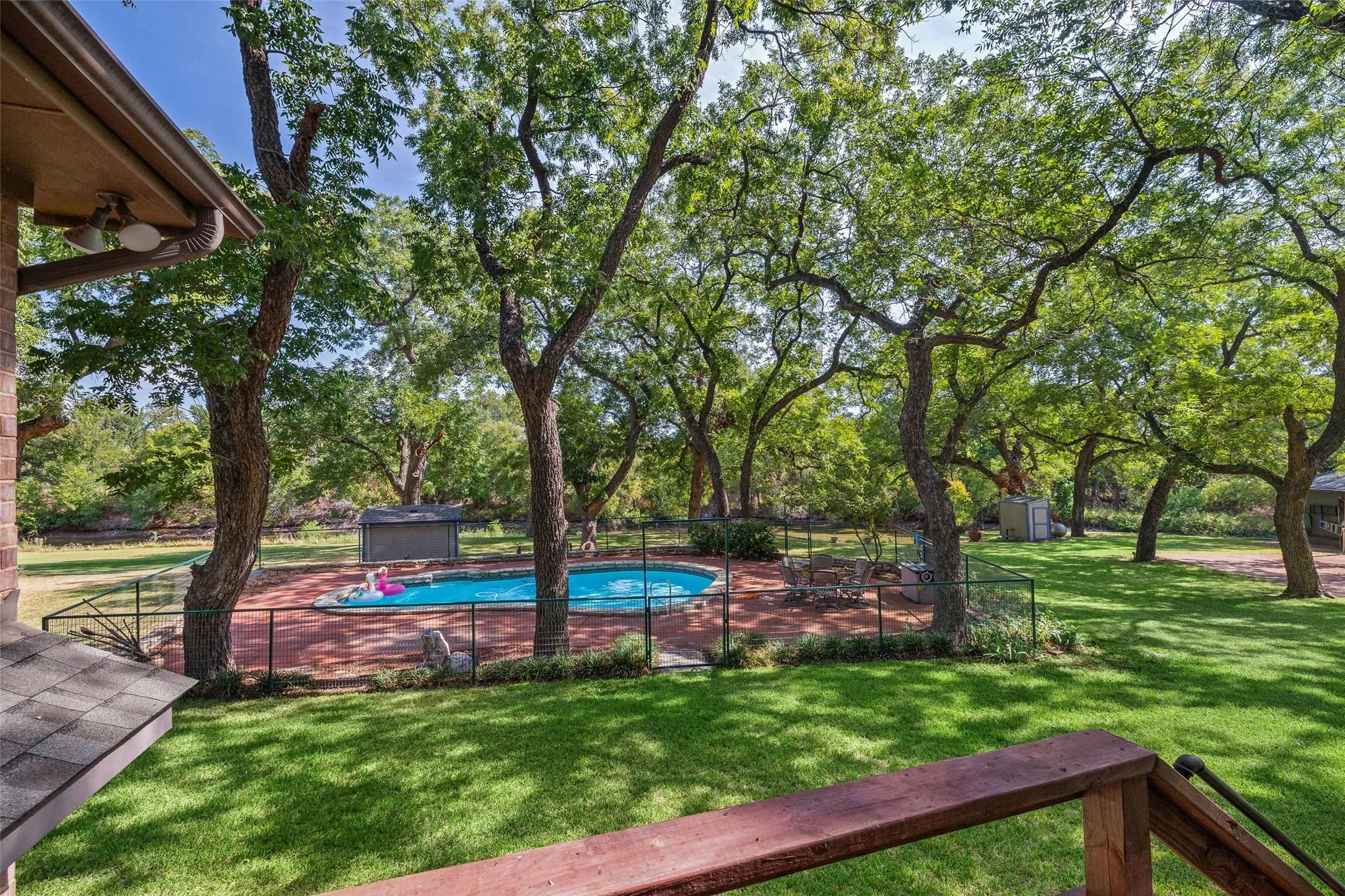 Expansive backyard with a fenced swimming pool, lush green lawn, mature trees, and a brick-paved pool deck
