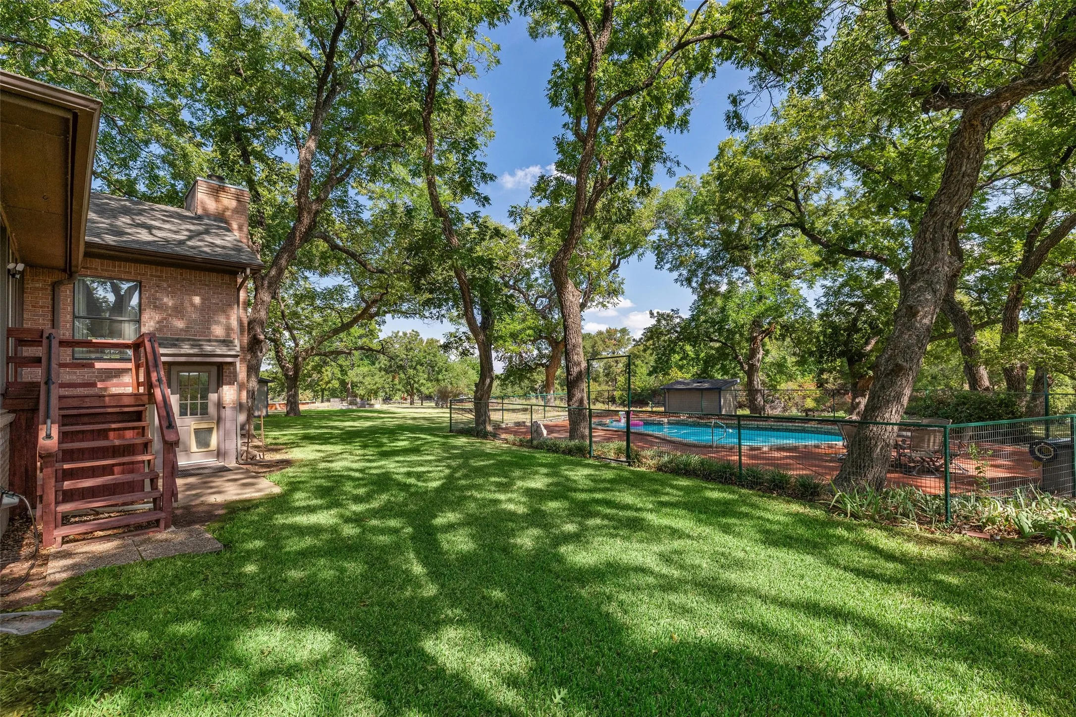 Expansive lawn with mature trees, a fenced swimming pool, and a brick-clad exterior with wood stairs