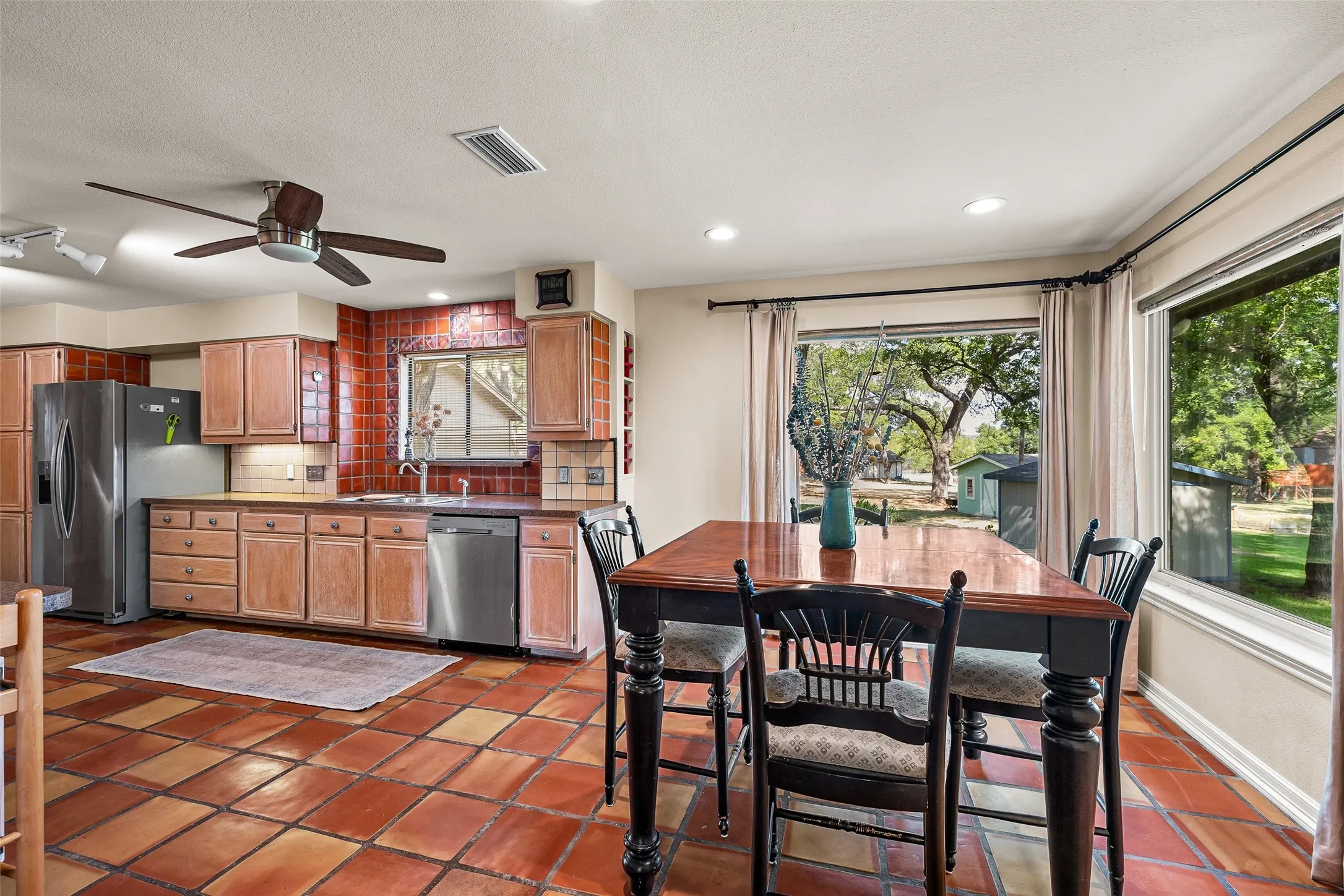 Kitchen and dining area featuring terracotta tile flooring, wood cabinetry, red square tile backsplash, stainless steel appliances, and large windows