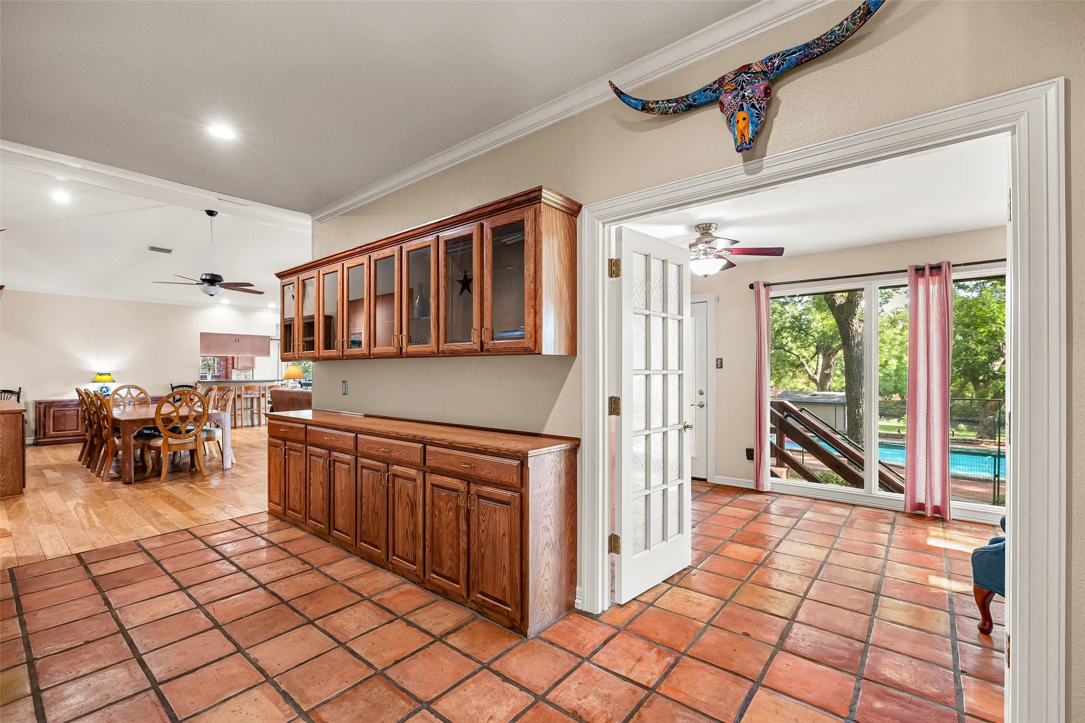 Terracotta tile flooring, built-in wooden cabinetry with glass-front uppers, French doors, and expansive windows offering a pool view