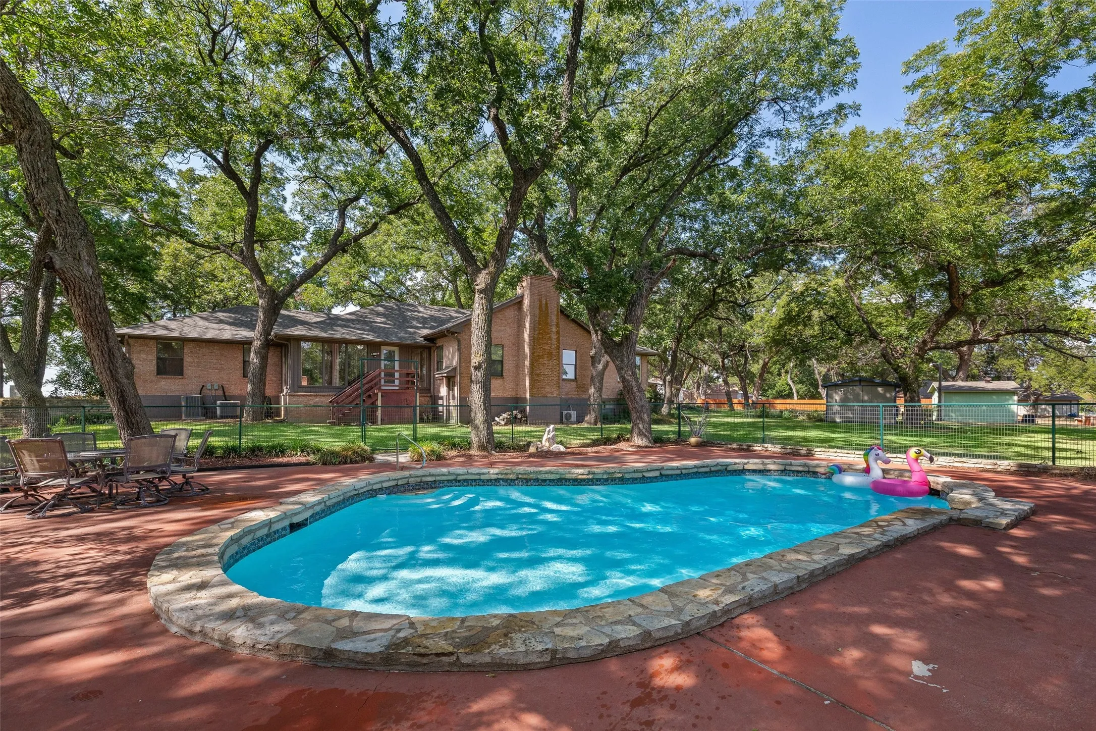 Curved in-ground pool with stone coping, surrounded by mature trees and a brick-clad residence