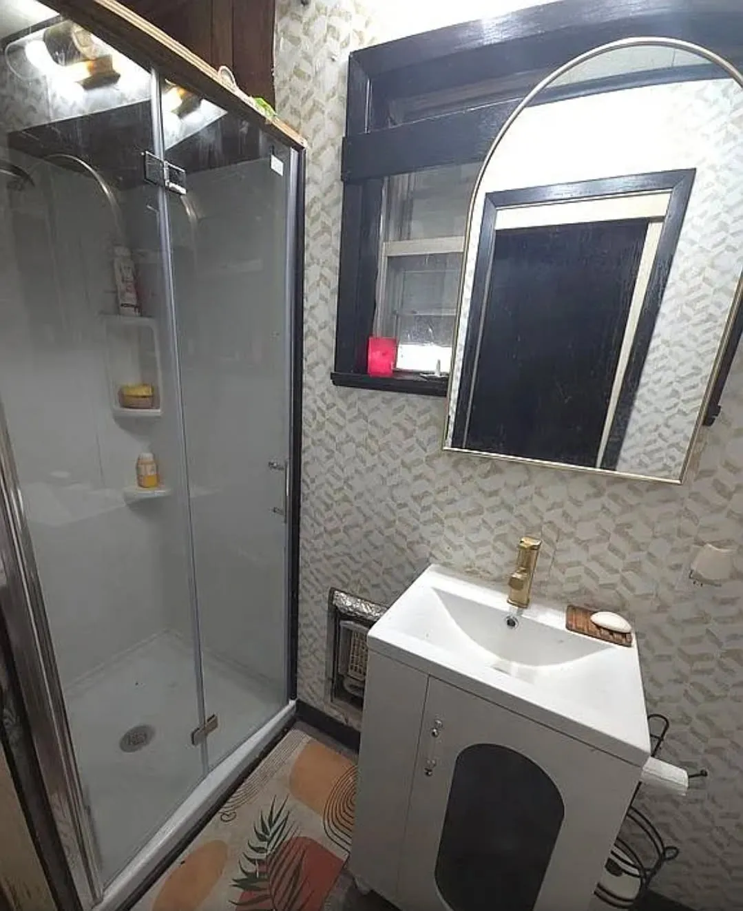 Enclosed shower with clear glass door, white vanity with integrated sink, gold-tone faucet, and arched wall mirror