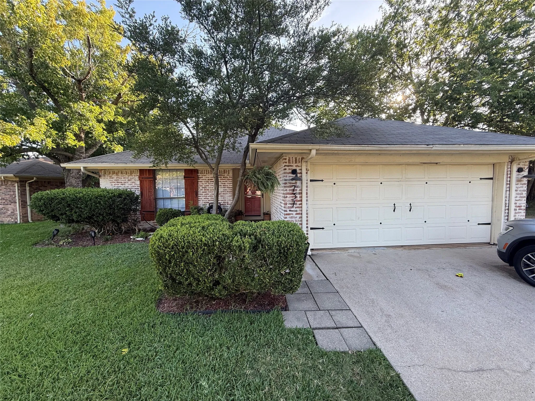 Brick facade with decorative shutters, attached garage, concrete driveway, and established front landscaping