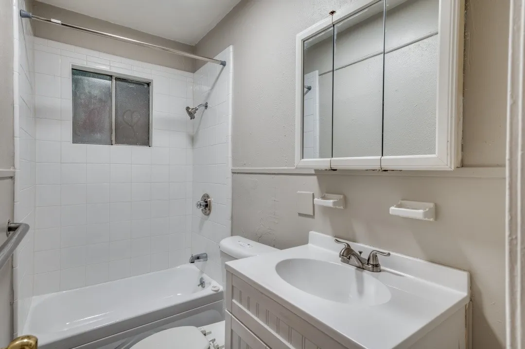 White tile shower surround with integrated window, white bathtub, white vanity with integrated sink, and mirrored medicine cabinet