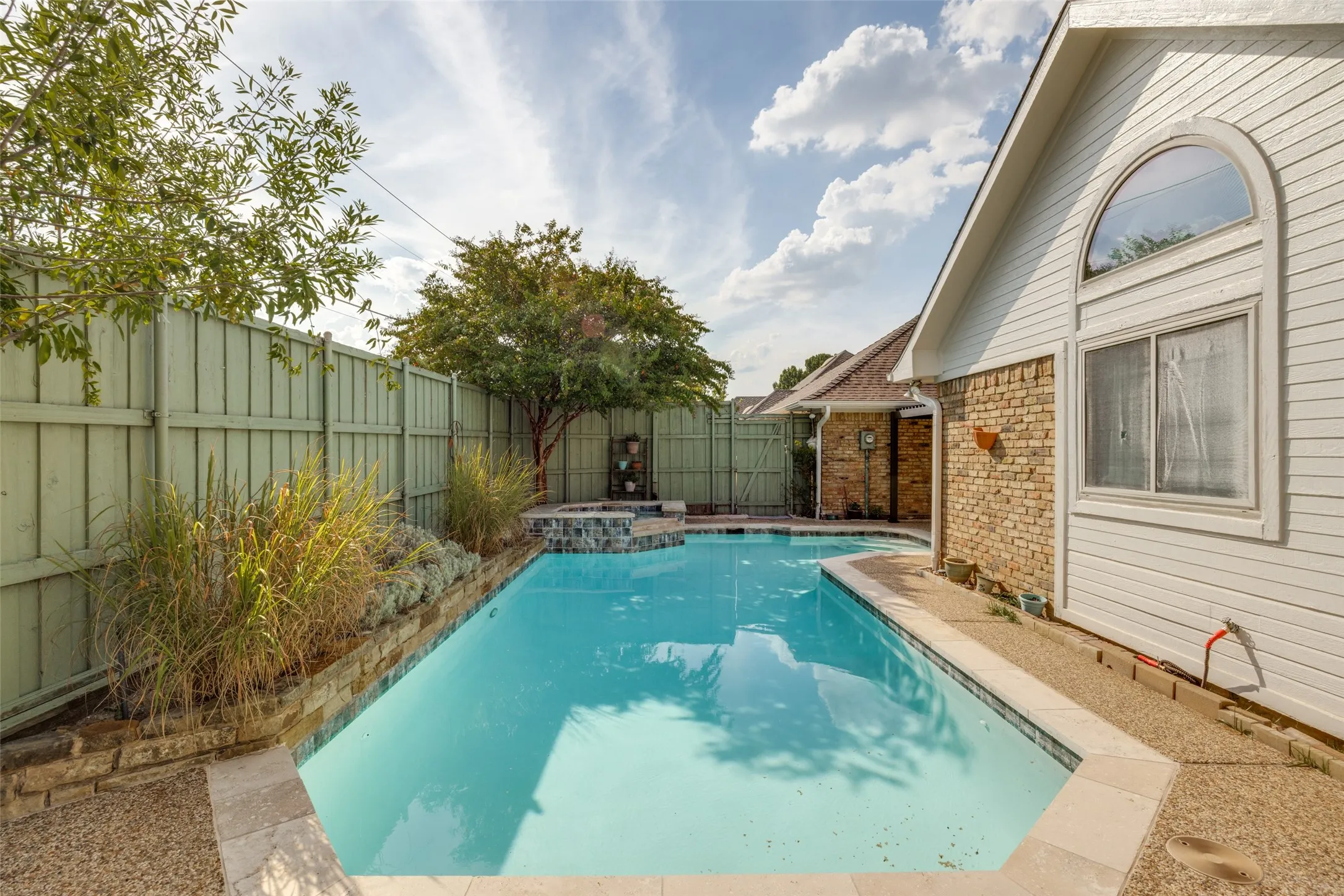 Outdoor swimming pool with integrated spa, bordered by a privacy fence and stone-finish retaining wall, adjacent to a structure featuring siding and brick accents