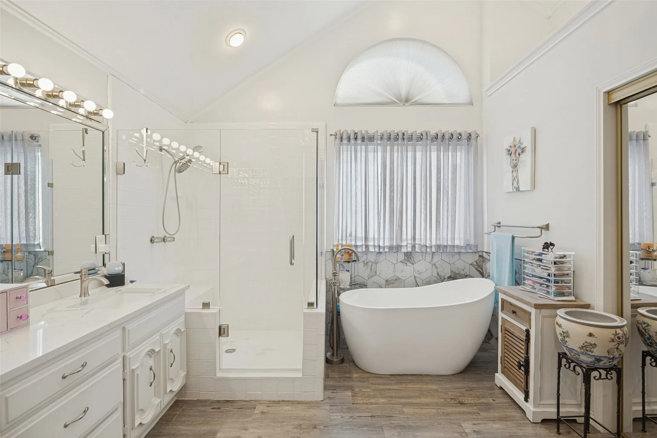 Vaulted ceiling with an arched window, freestanding soaking tub, glass-enclosed shower, dual vanity with undermount sinks, and wood-finish flooring