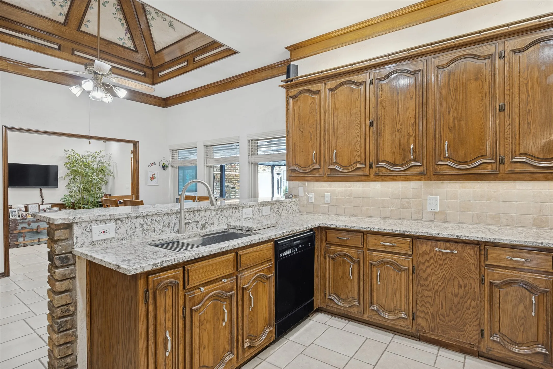 Kitchen featuring wood cabinetry, farm sink, granite countertops, tile flooring, a tile backsplash, and a coffered ceiling with a central fan