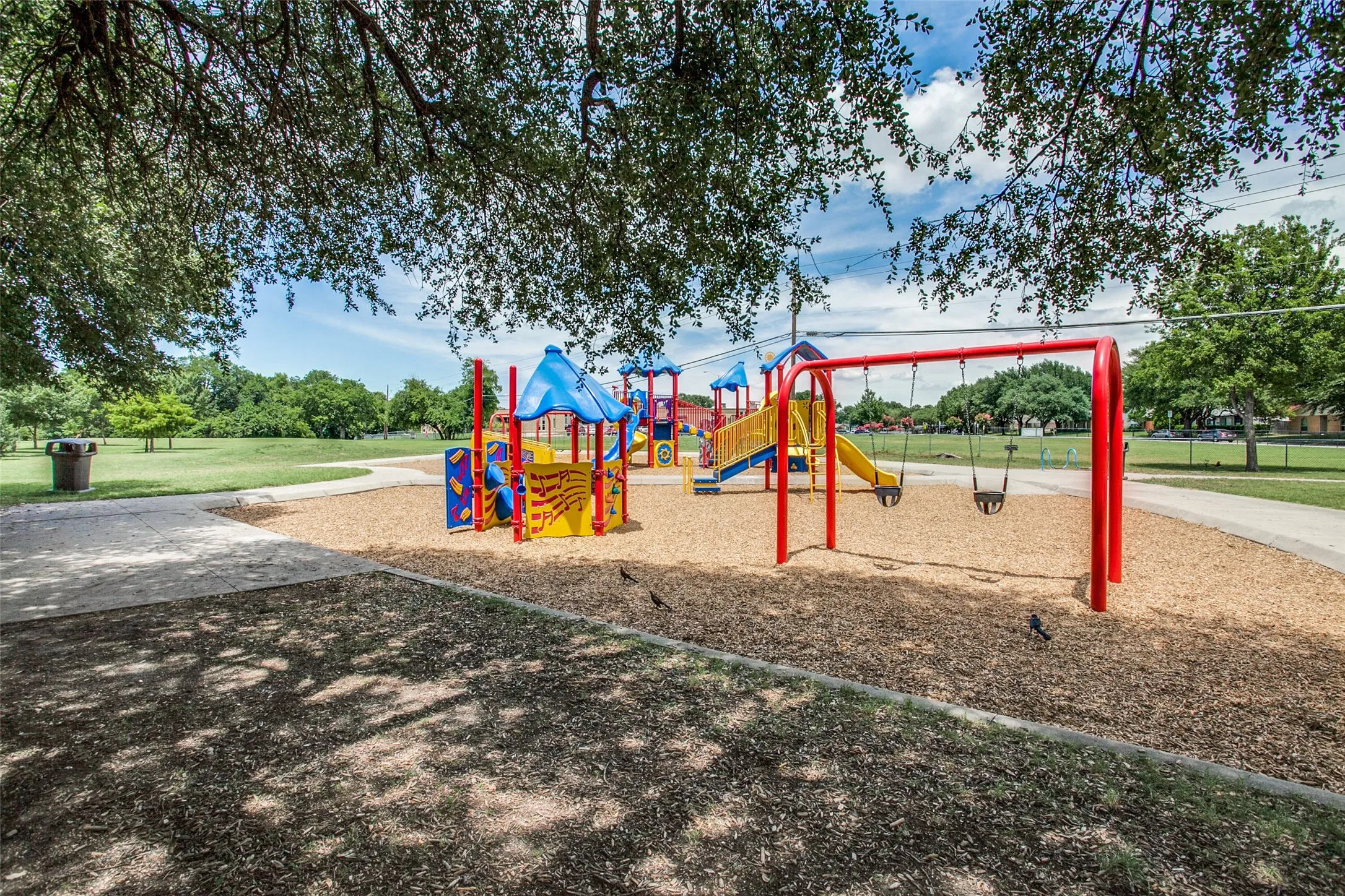 Recreational playground with colorful equipment, swings, and a wood chip surface, bordered by a paved pathway and mature trees