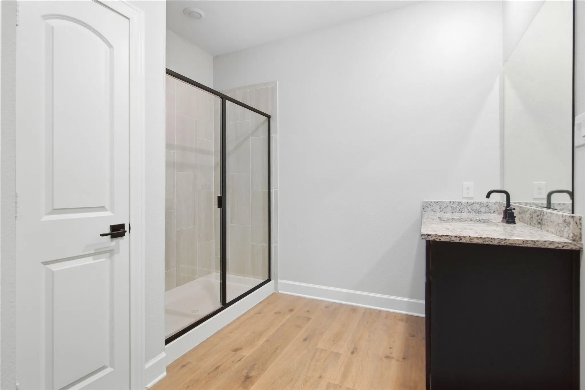 Black-framed glass shower enclosure, single vanity with granite countertop, matte black faucet, and wood-finish flooring