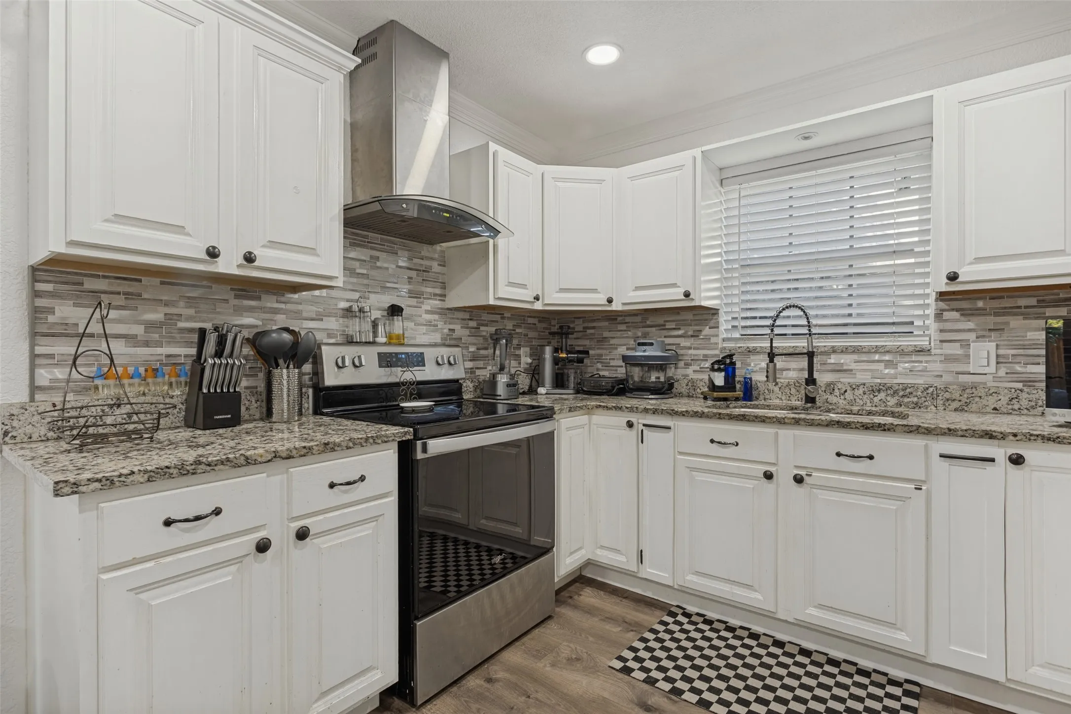 Kitchen featuring white shaker-style cabinetry, stone-finish countertops, linear mosaic tile backsplash, stainless steel range with overhead chimney vent, and wood-finish flooring