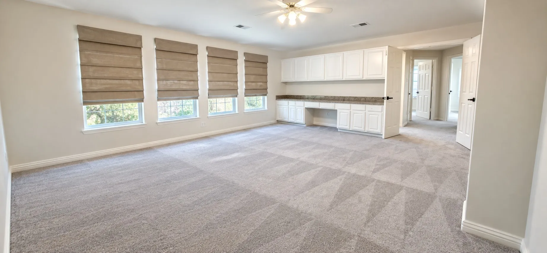 Spacious room featuring light-colored carpeting, multiple windows with neutral roman shades, and a built-in desk with cabinetry